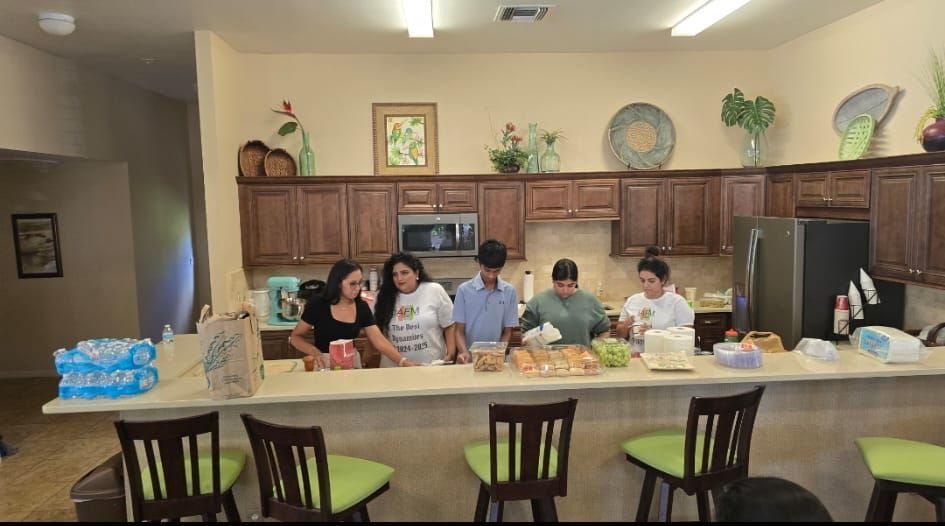 People preparing food at a kitchen counter. Brown cabinets, bar stools, and a refrigerator.