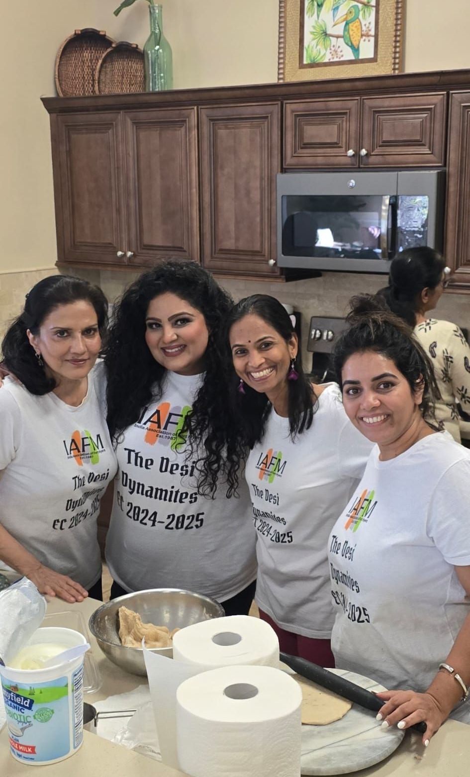 Four women in matching t-shirts smile in a kitchen. They are preparing food; there are ingredients and a rolling pin.