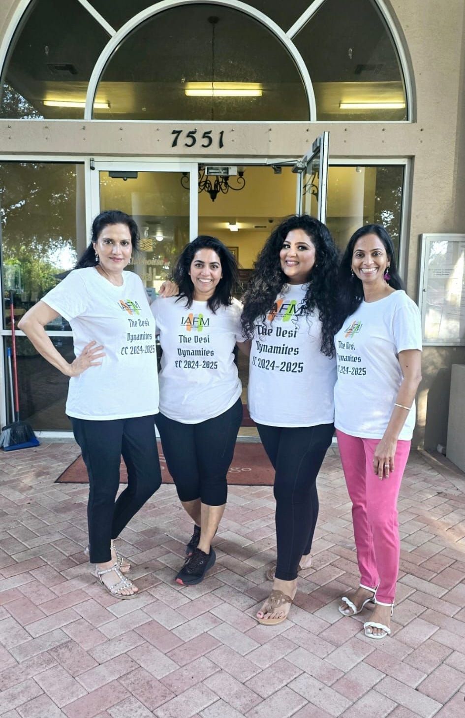 Four women in white tees and leggings pose in front of a building with 