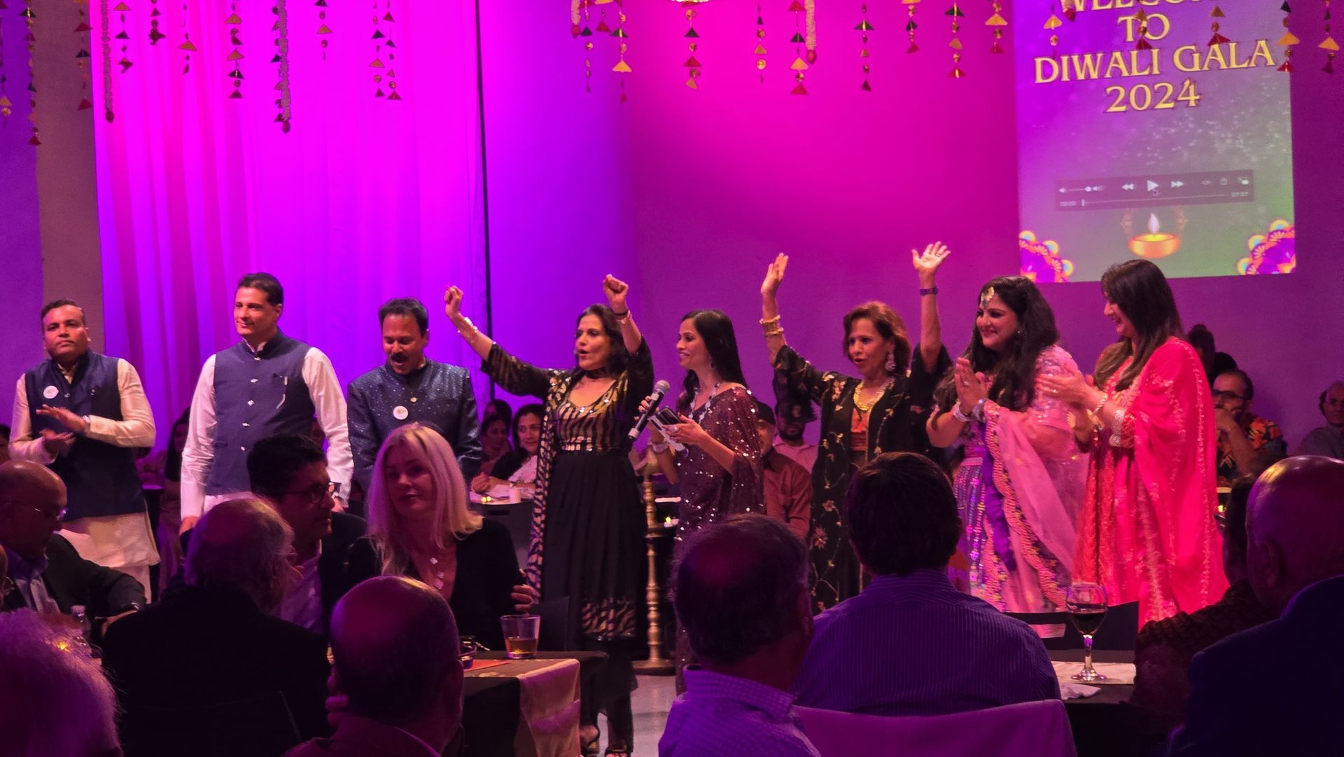 People on stage at a Diwali gala, dancing and waving, with an audience below, lit in pink and purple.