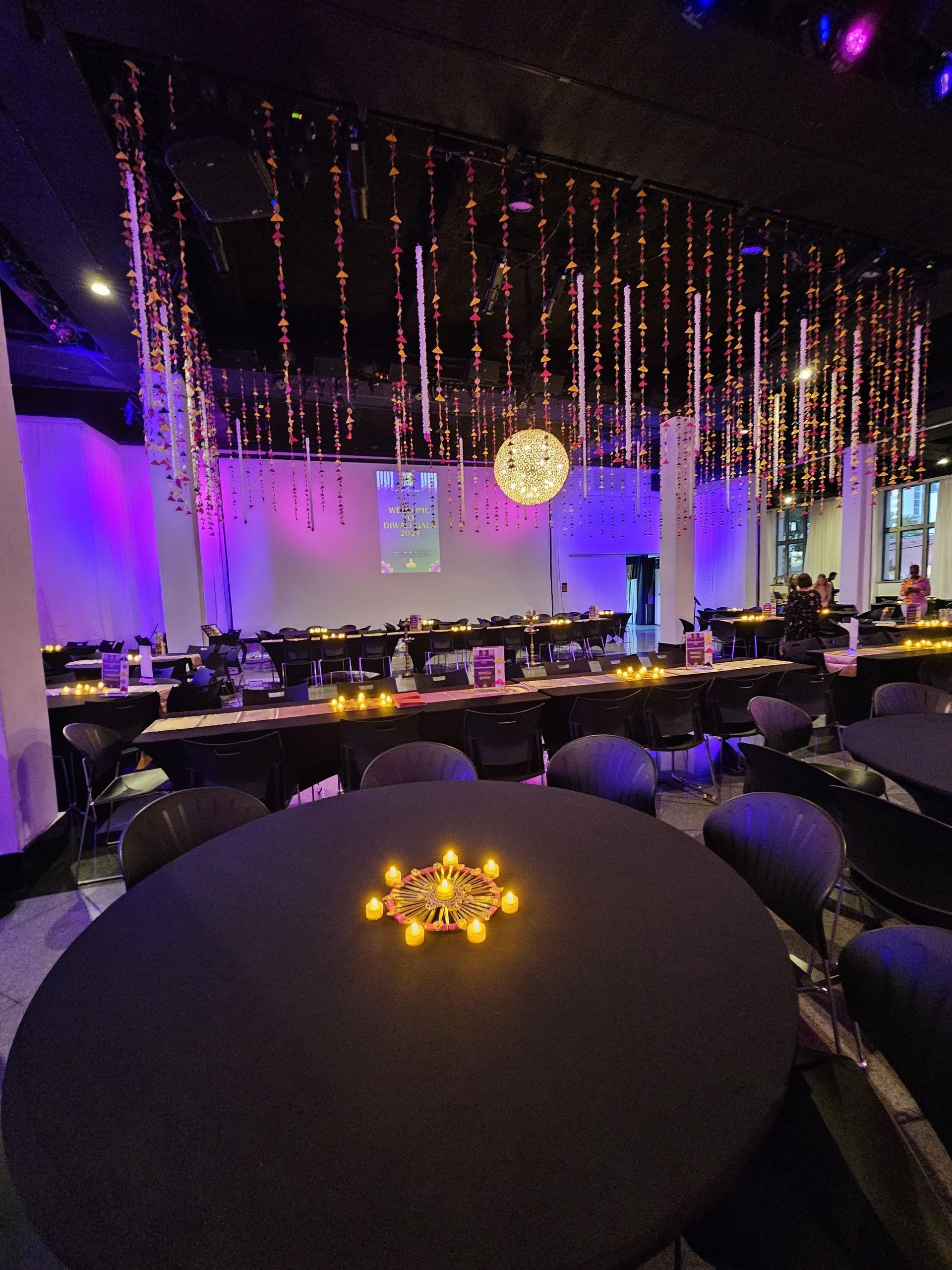 Indoor event space, round black tables set for dining, decorated with candles and hanging lights.