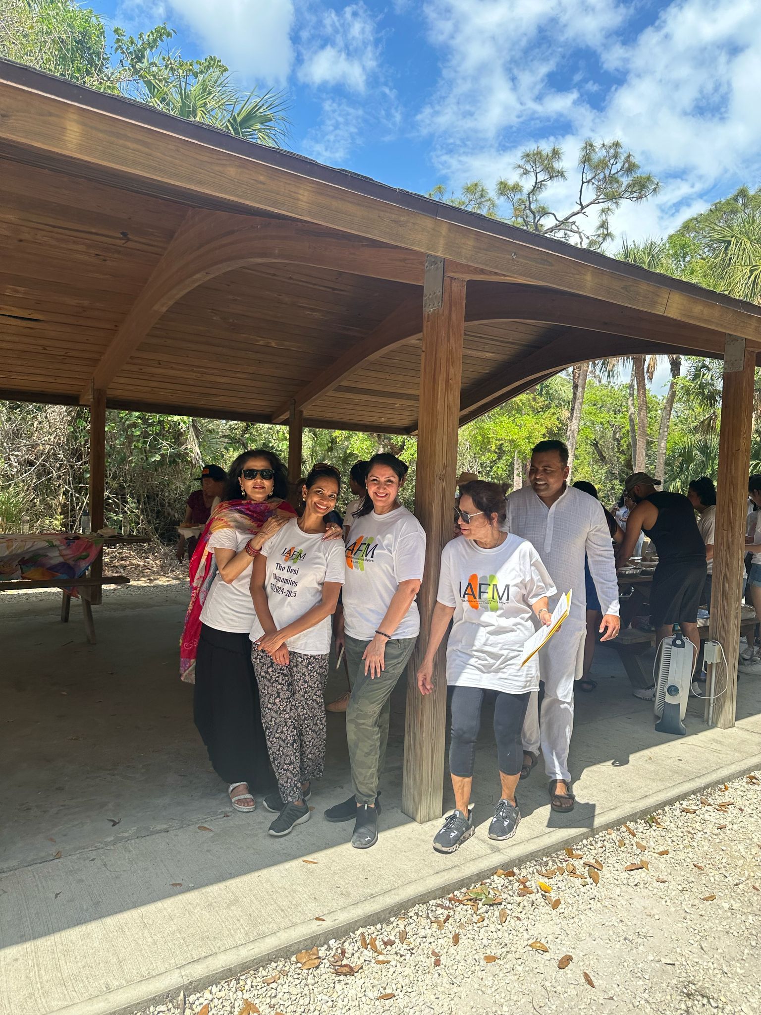 Group of people under a wooden shelter in park. People in white shirts smile, some look at camera.
