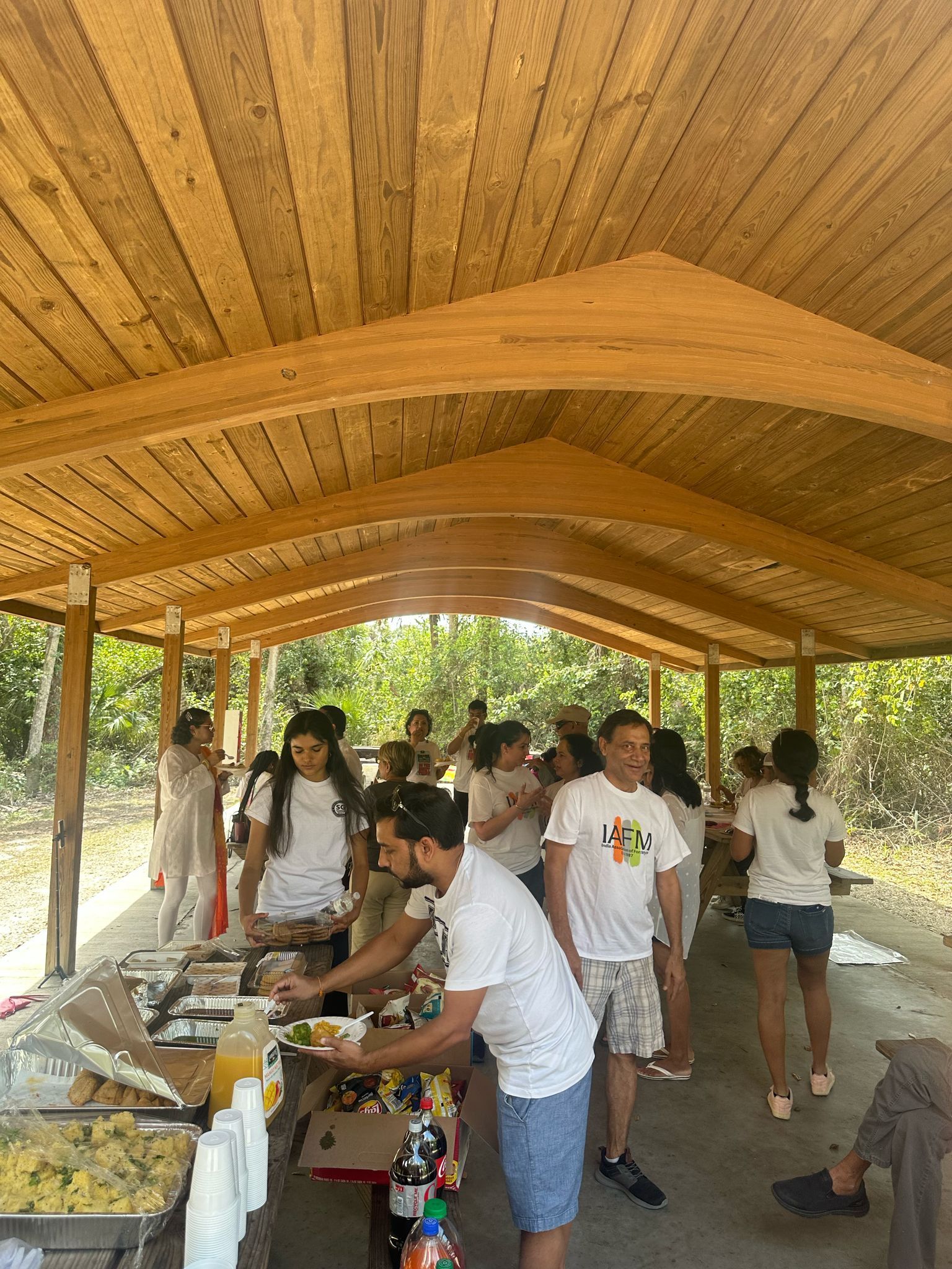 People gathered at a picnic under a wooden shelter; food is on a table.