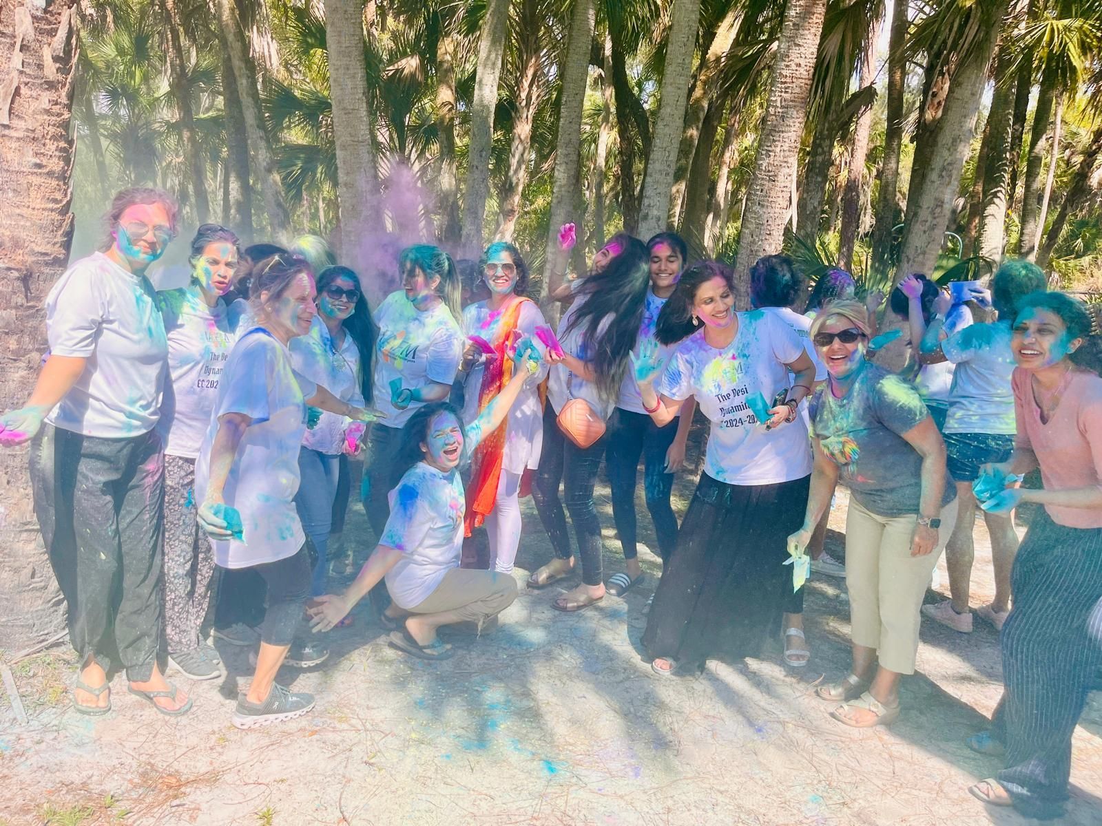 Group of women in white shirts covered in colorful powder, celebrating outdoors among palm trees.