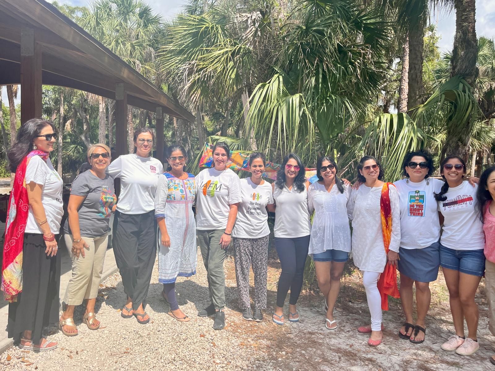 Group of women smiling at a park; trees in the background. They are wearing white tops and some colorful accents.