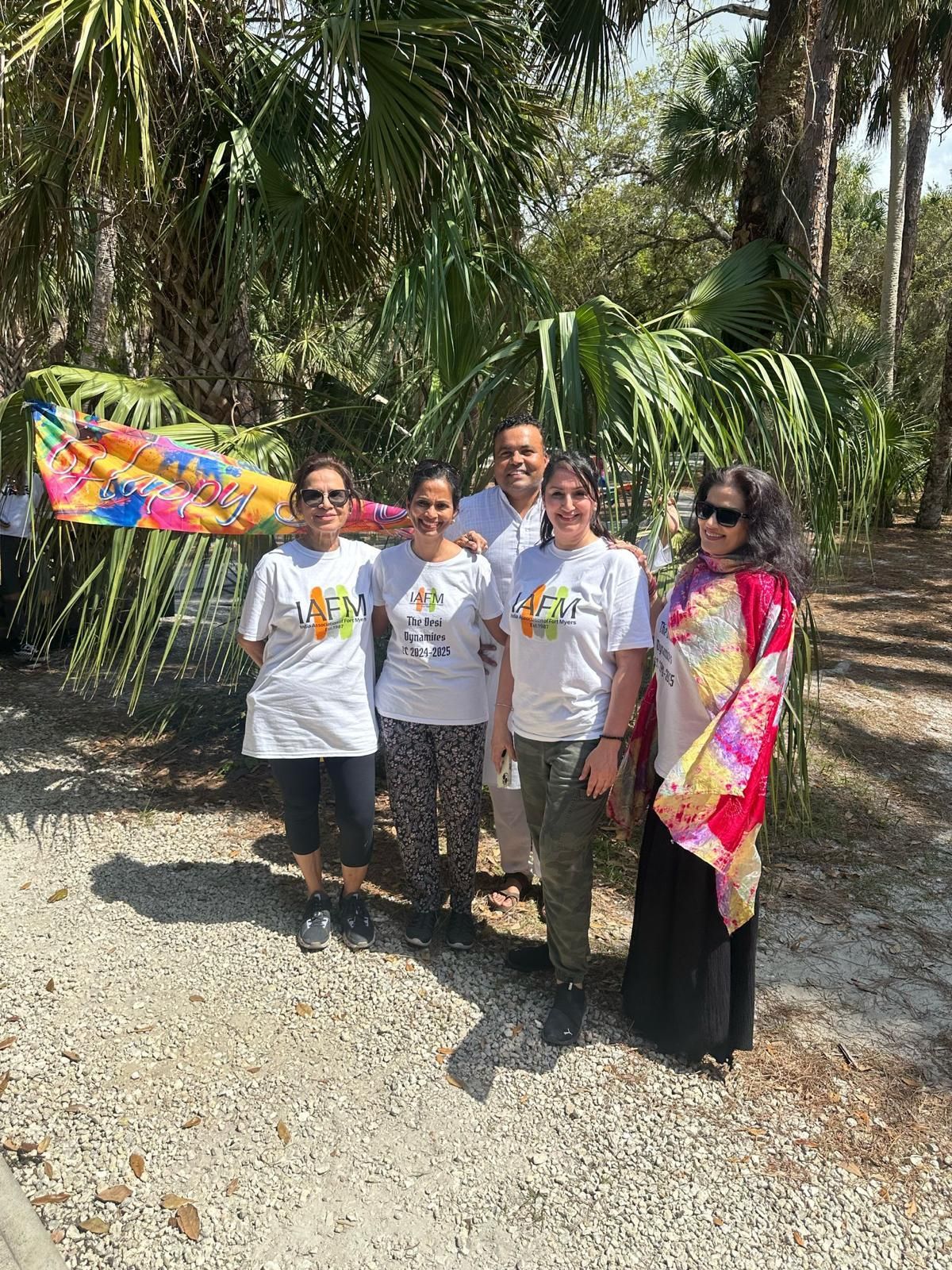Group of five people posing outdoors, wearing white shirts with a colorful banner in the background.