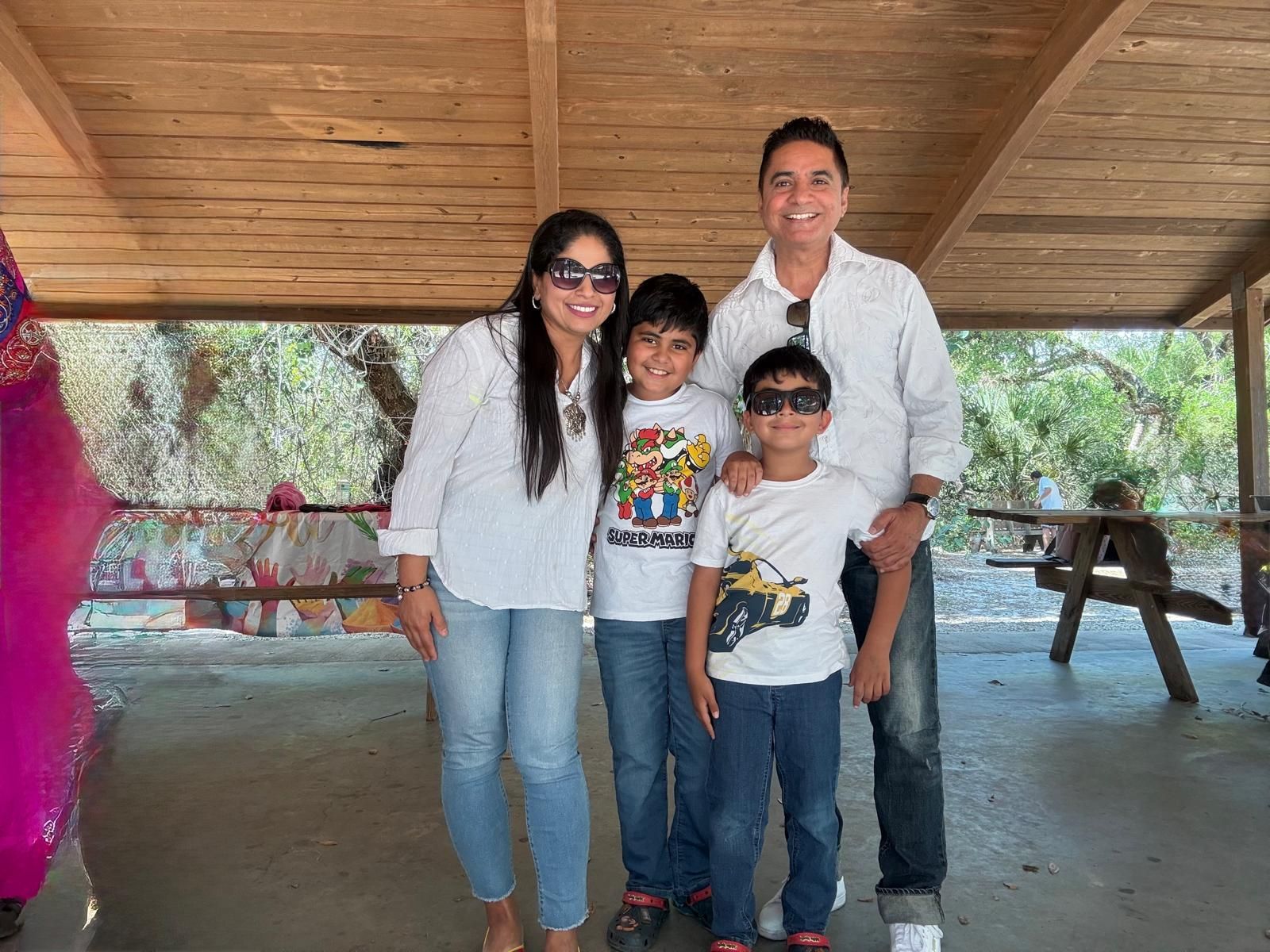 Family of four smiling in a shaded outdoor area; they wear sunglasses and casual white shirts with jeans.