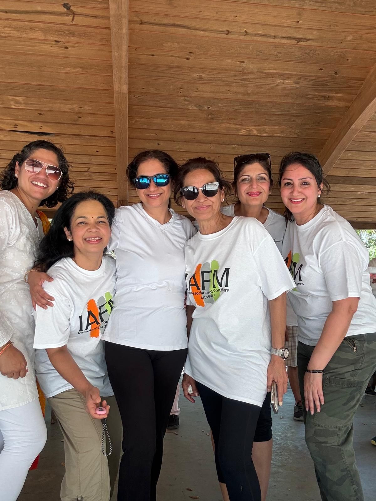 Six women smiling, wearing white t-shirts with a logo, posing outdoors under a wooden shelter.