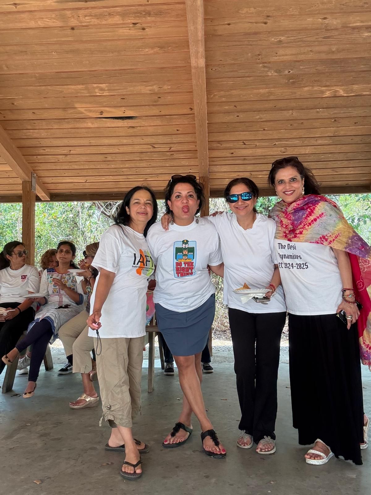 Four women pose together, smiling, under a wooden shelter. Others sit in the background.