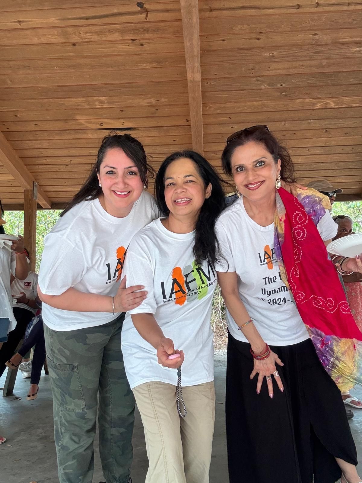 Three women smiling at the camera, wearing matching white t-shirts. Outdoors under a wood-paneled roof.