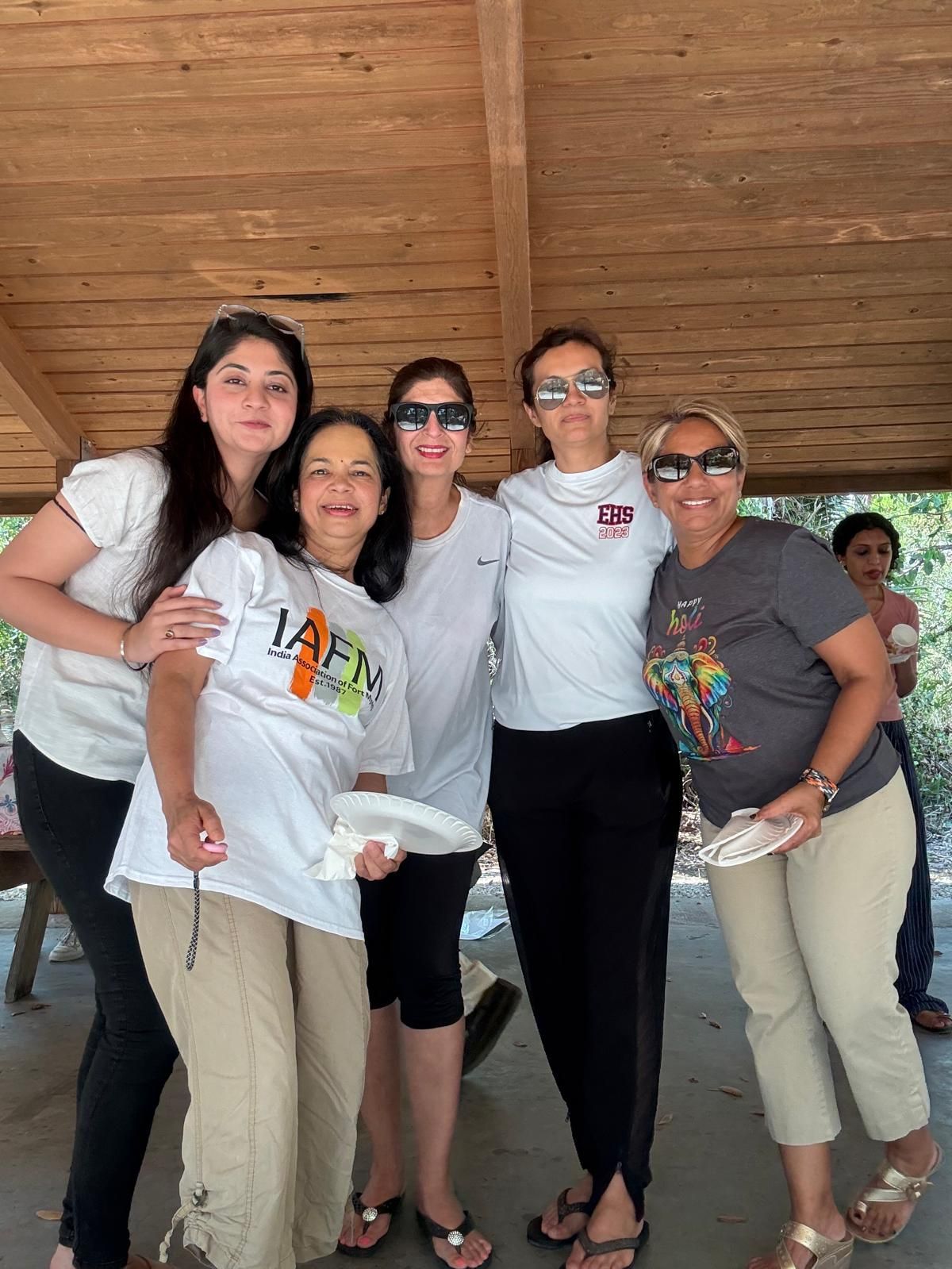 Five women pose under a wooden shelter. Two wear white shirts. All are smiling.