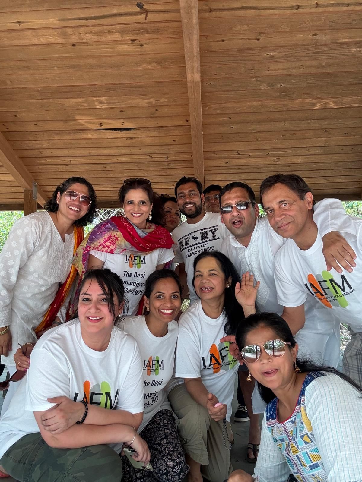 Group of people wearing white shirts, smiling, and posing under a wooden structure.