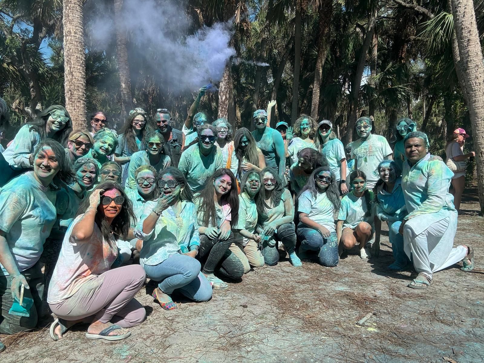 Group of people covered in blue powder outdoors, posing for photo; trees in background.