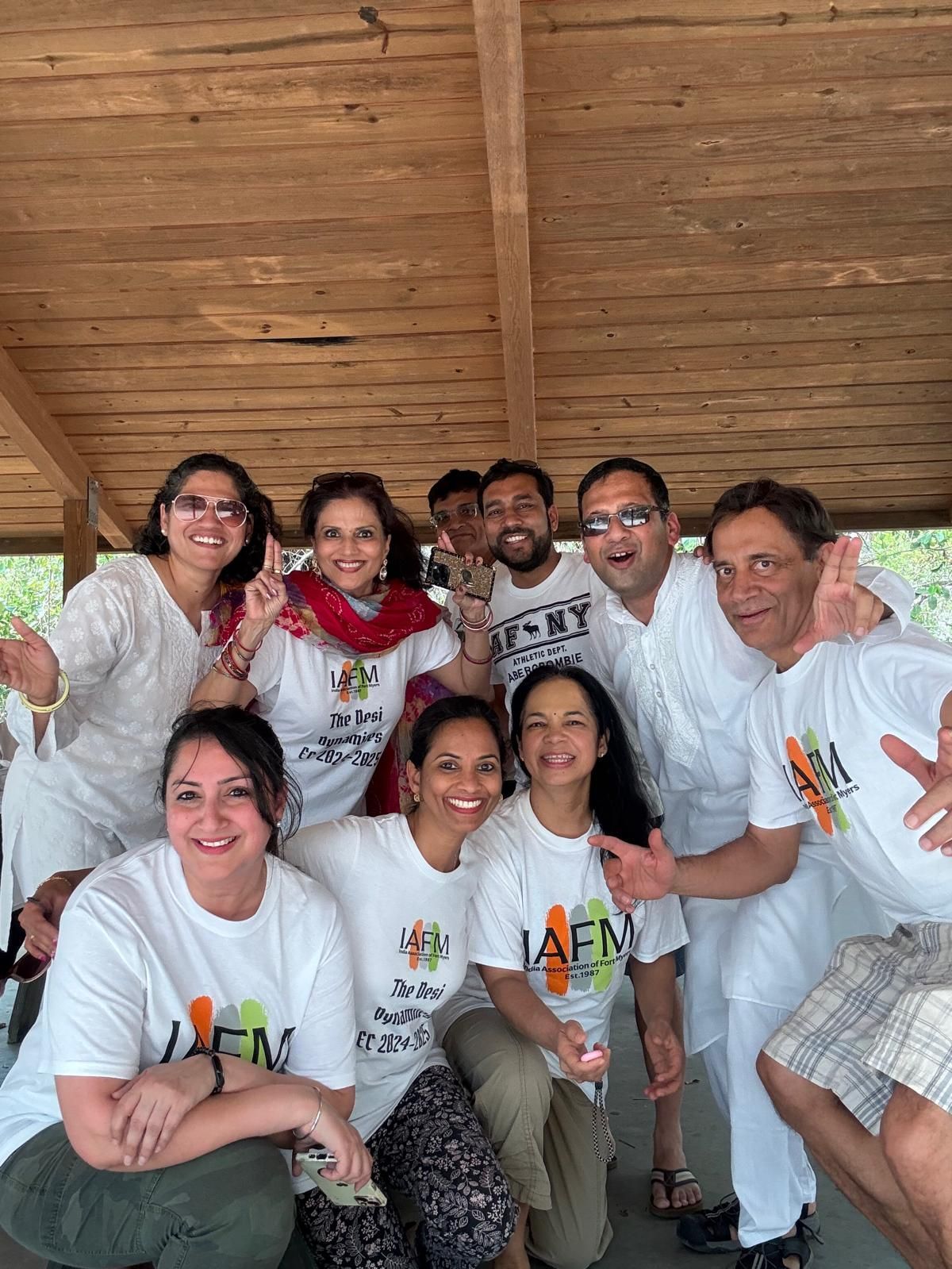 Group of people smiling, posing under a wooden structure, wearing white t-shirts, some with colorful accents.
