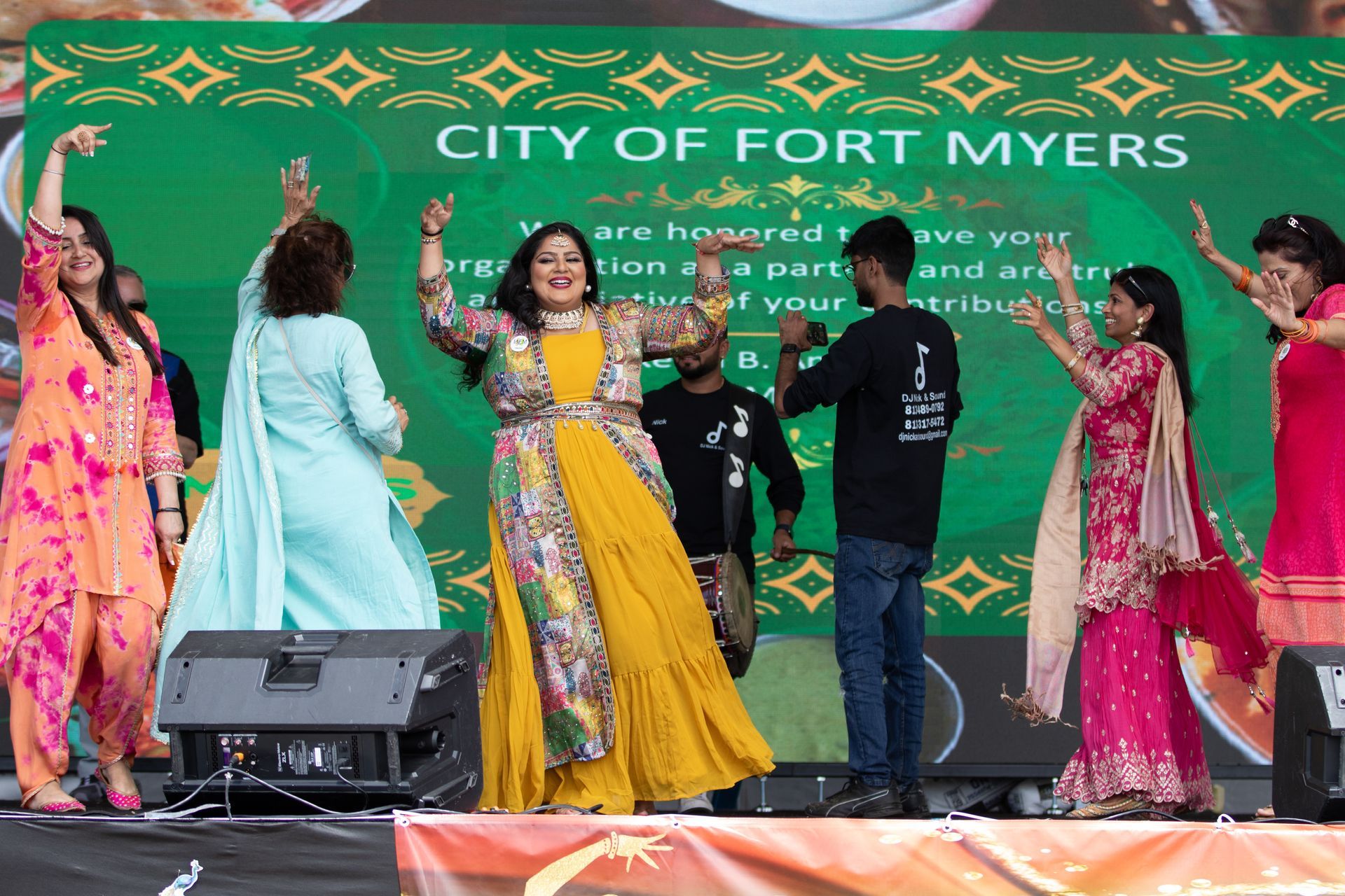 People dancing on stage at a Fort Myers city event. Bright clothes, green screen, and a crowd.