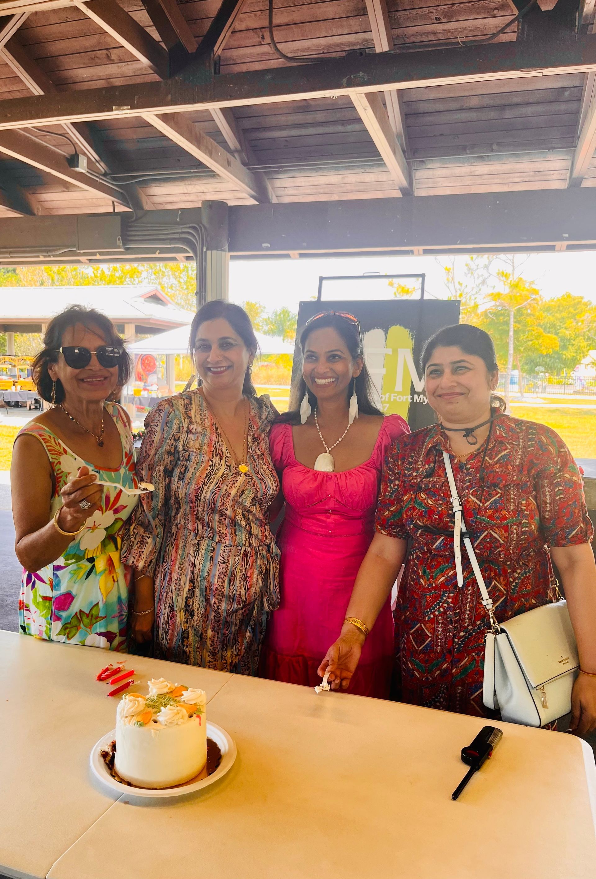 Four women smiling near a table with cake, under a park shelter.