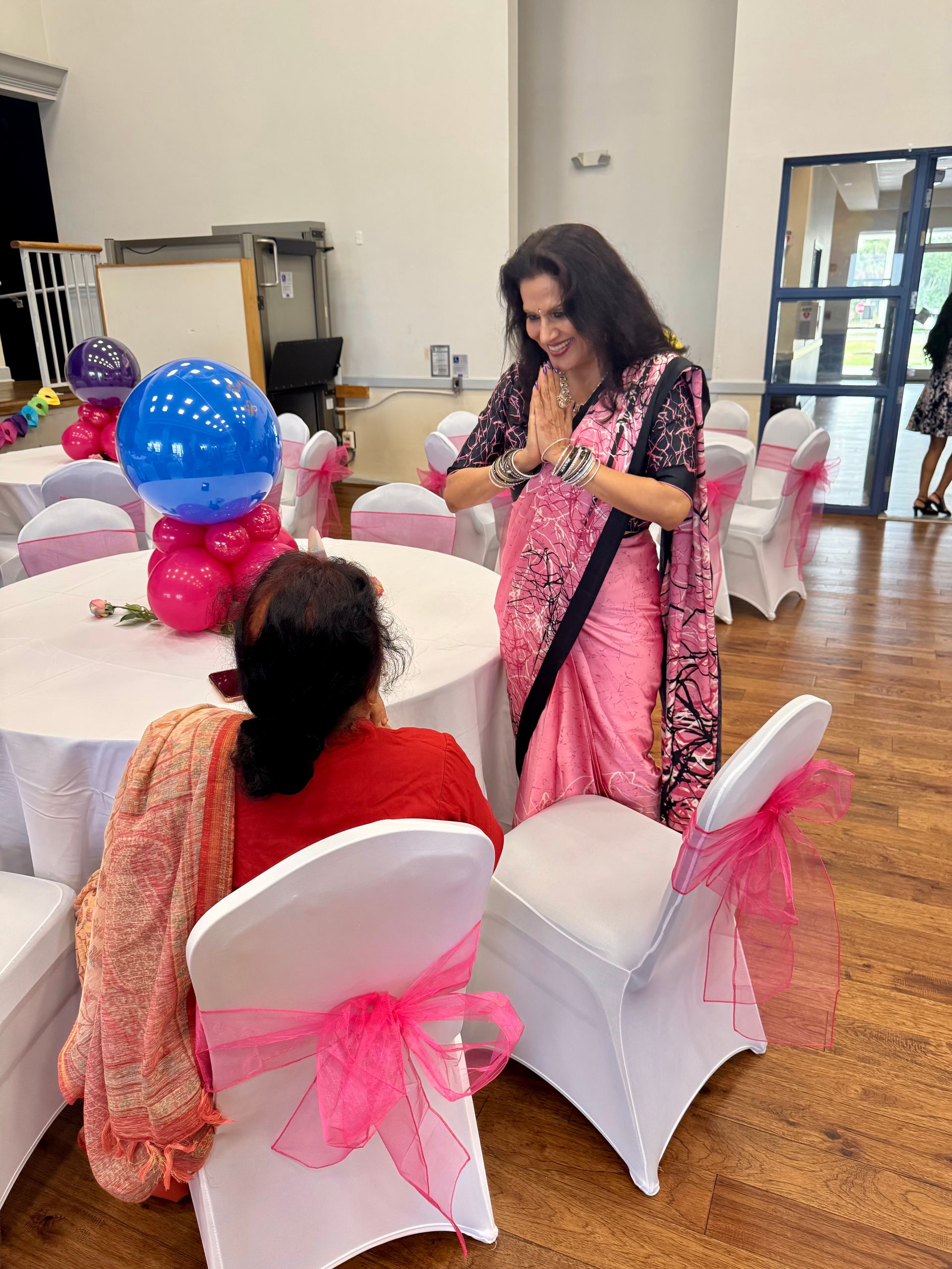 Woman in pink sari greeting seated guest at event with decorated tables and chairs.