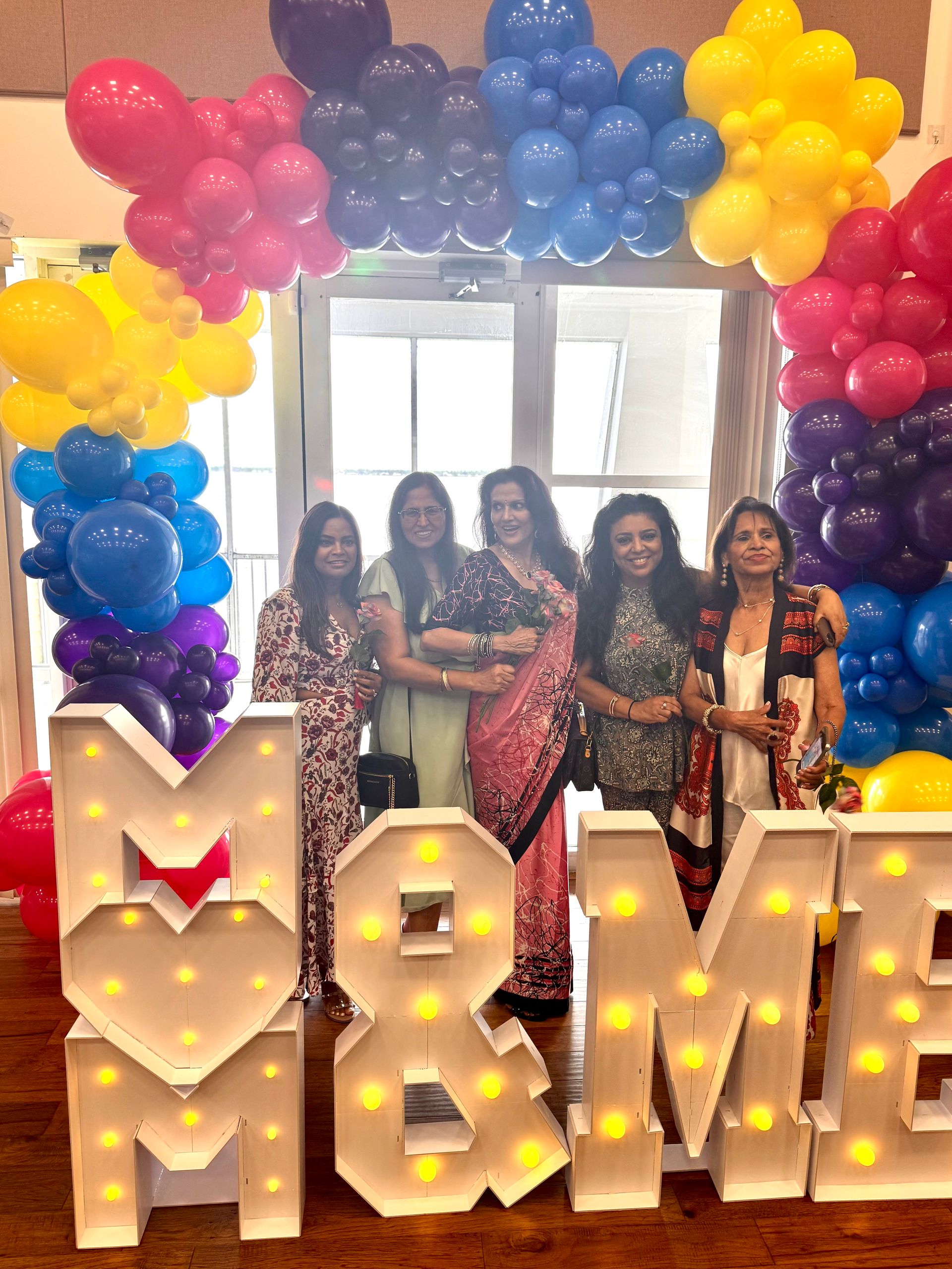 Five women pose in front of a balloon arch and light-up letters that read 