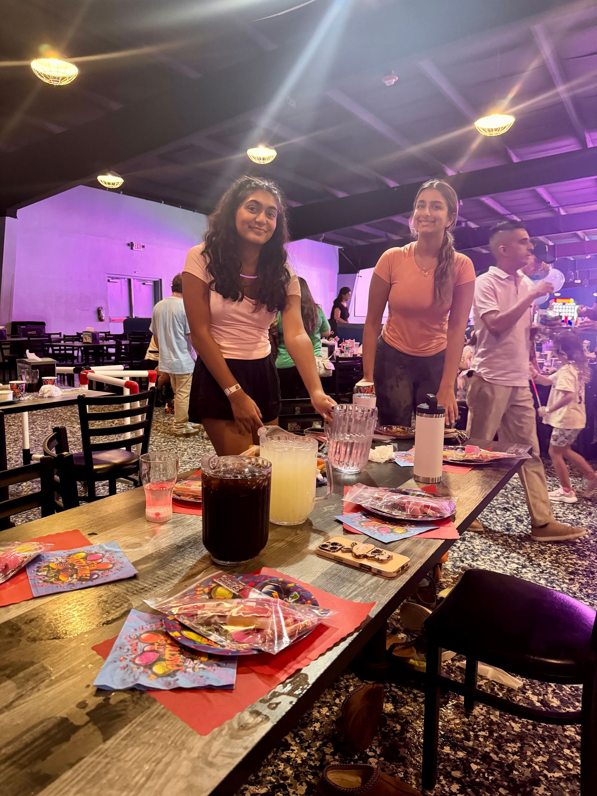 Two women at a bar with drinks, smiling. Colorful plates on the counter, and other people are in the background.
