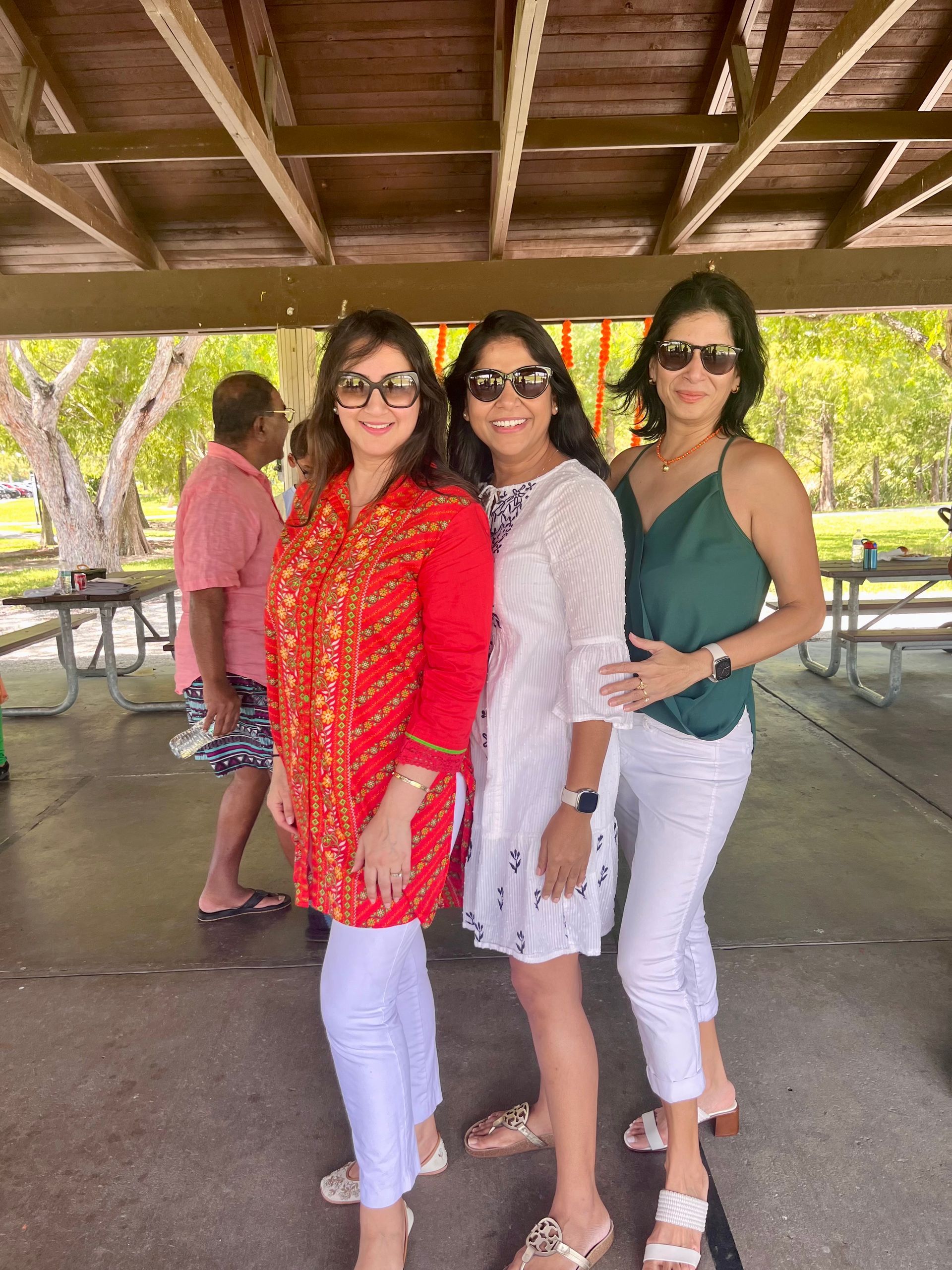 Three women smiling at a park picnic. They wear colorful outfits. Man in pink in background.