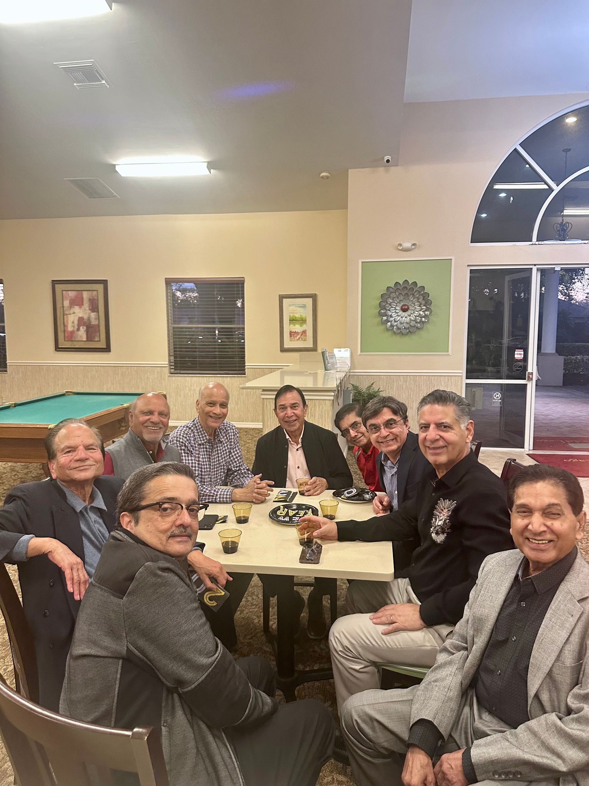 Group of men seated around a table, possibly playing cards, in a room with a pool table and arched doorway.