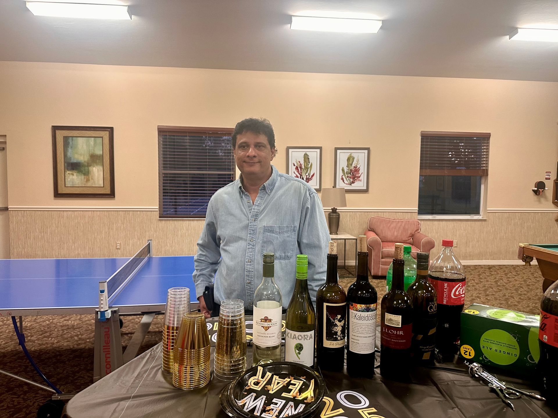 Man stands behind a table with drinks and decorations in a room with a ping pong table.