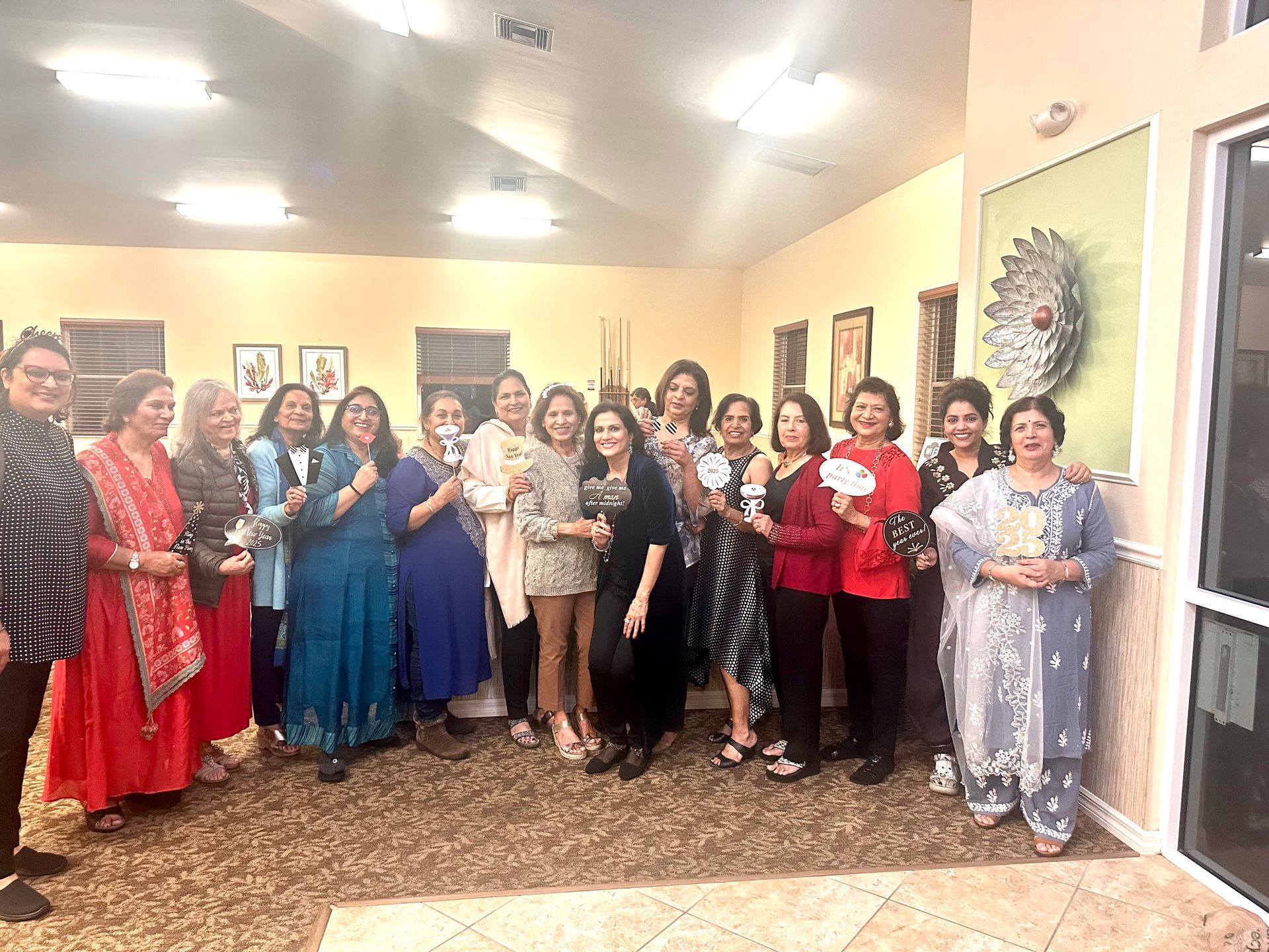 Group of women in a well-lit room, posing for photo; many smiling, some holding items, art on walls.
