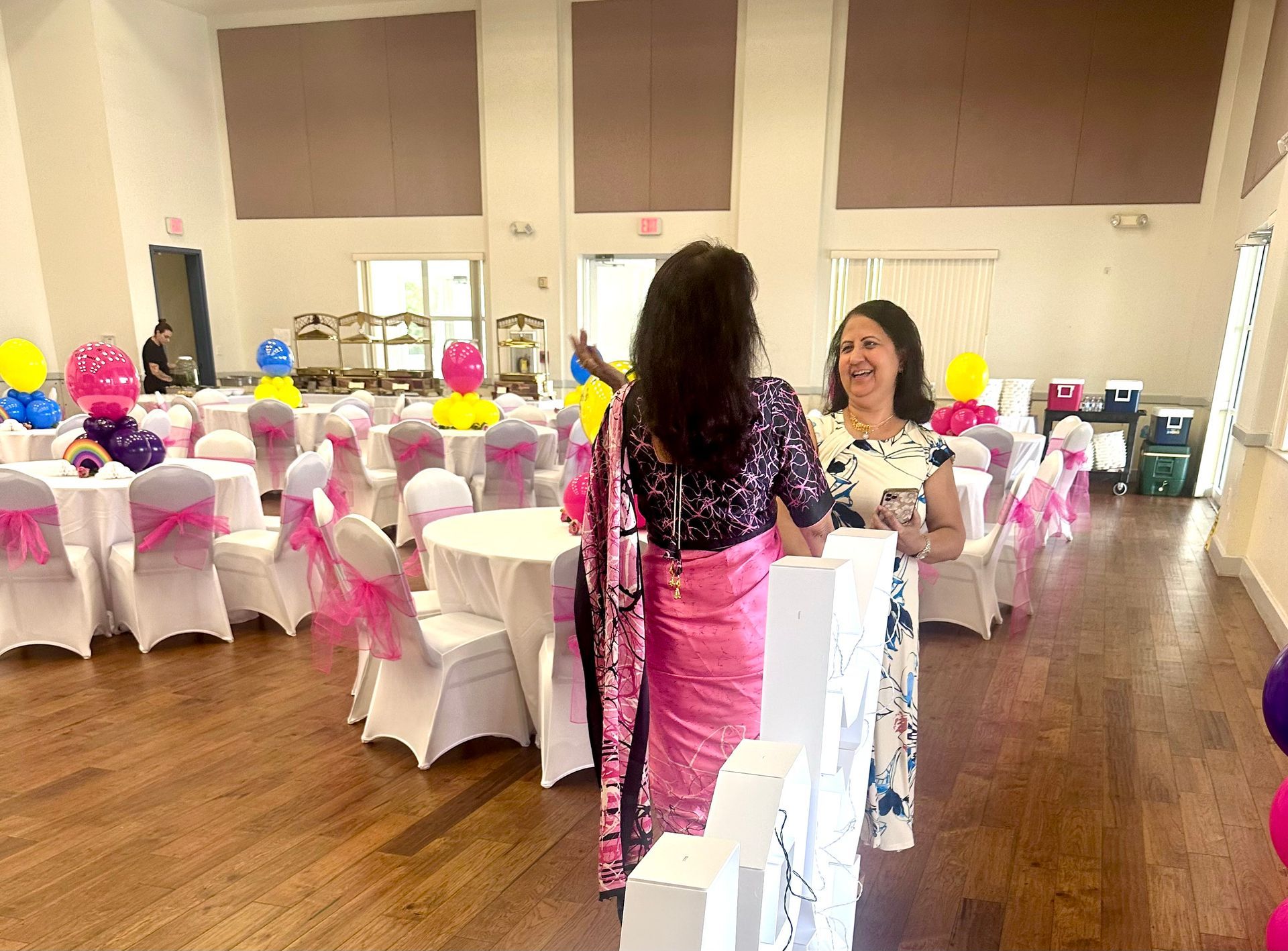 Two women at party, one in pink sari, other in floral dress, next to decorated tables.