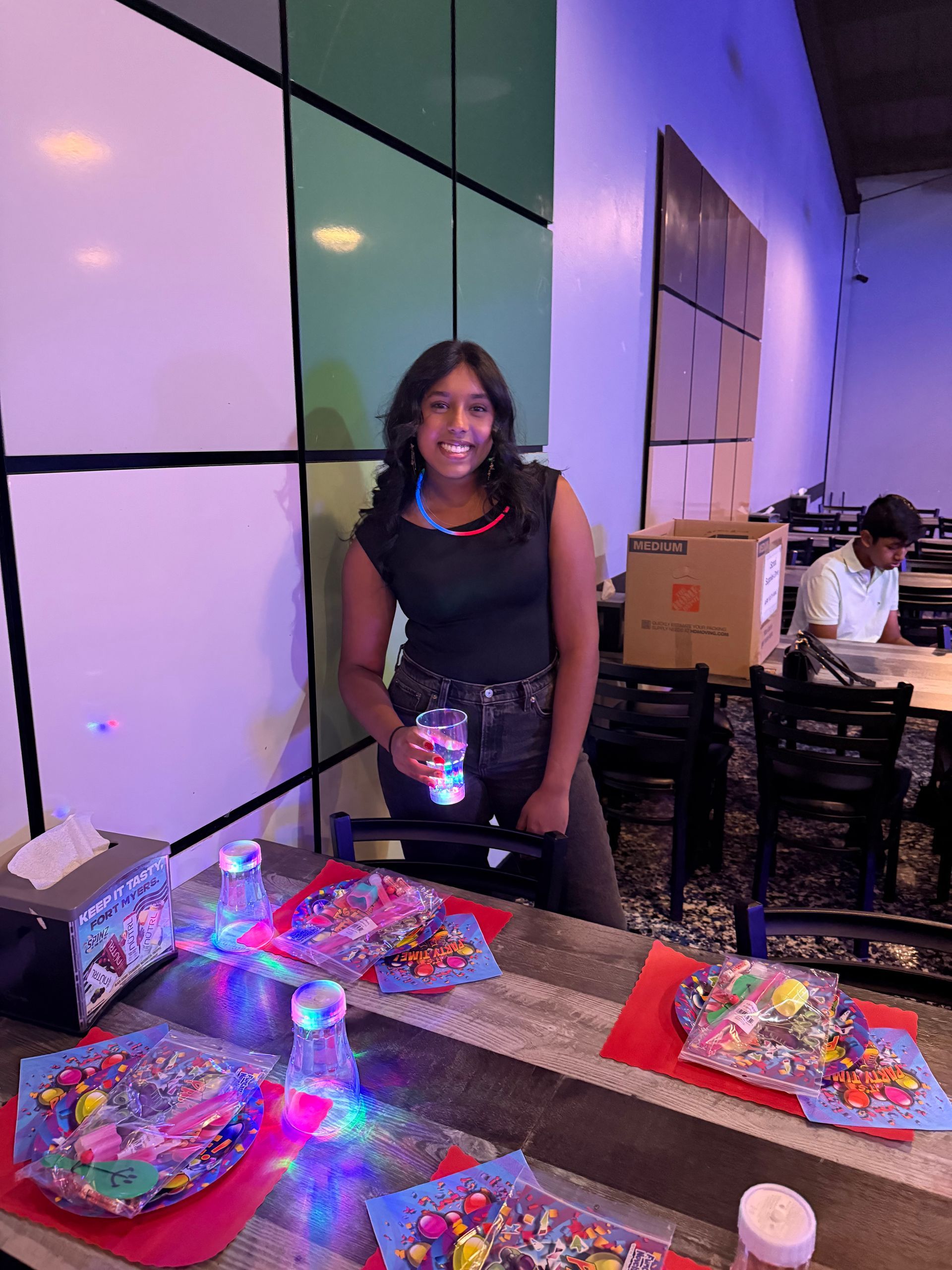 Woman smiling at a table with crafts, inside a restaurant. She's holding a drink. The setting is well-lit.