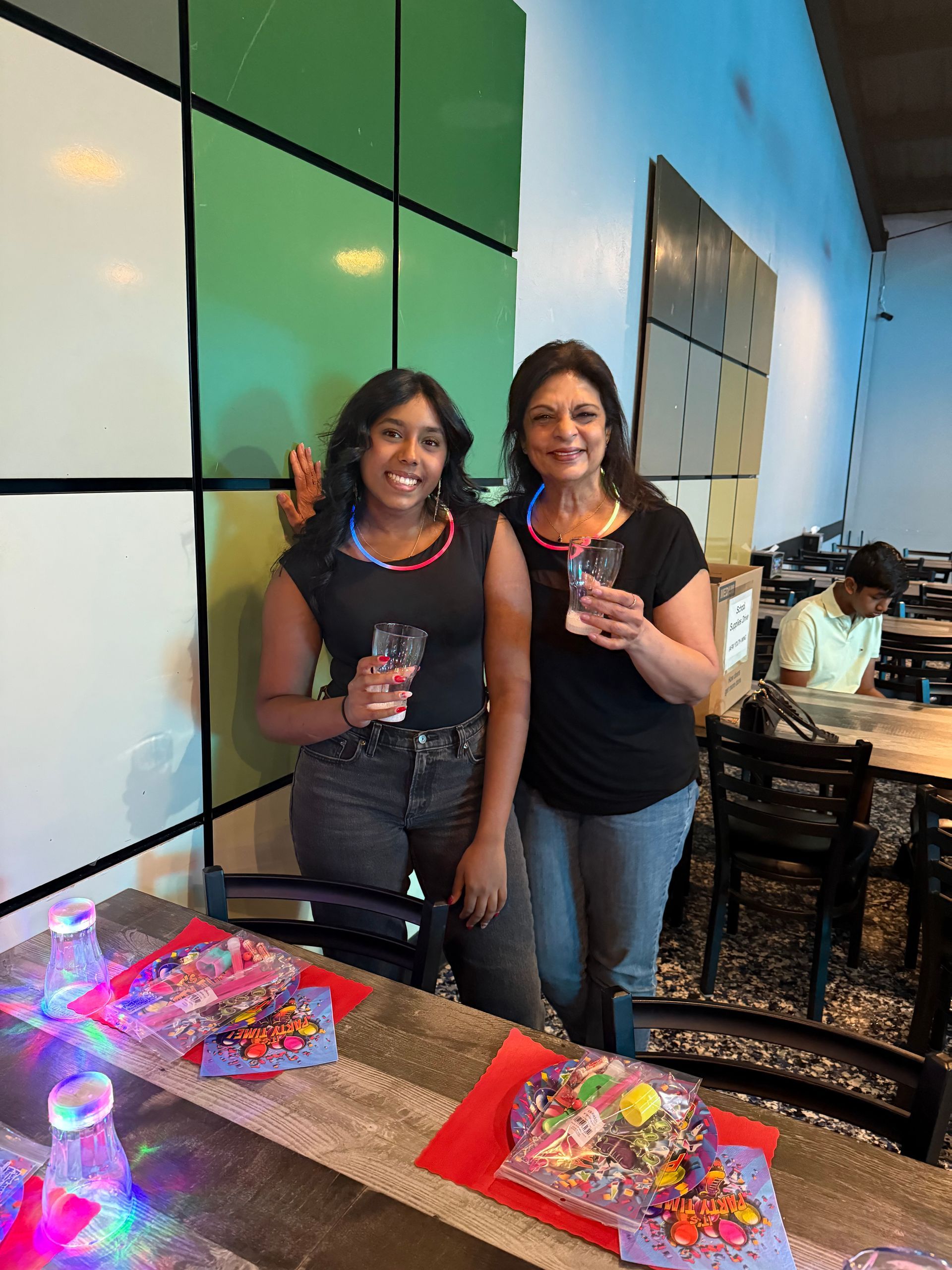 Two women with drinks pose at a table in a brightly colored venue.