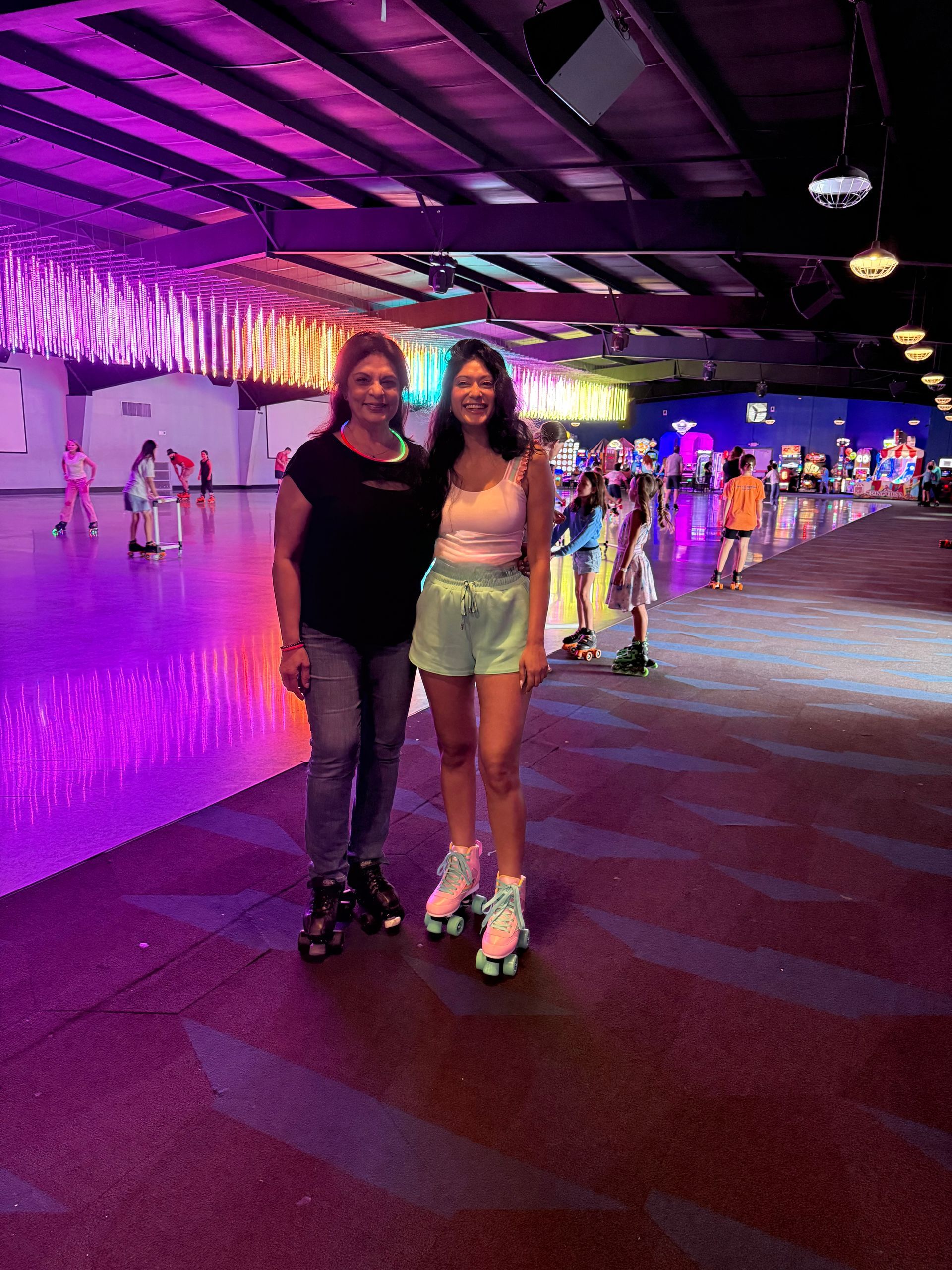 Two women pose at a roller rink, wearing skates. The room is brightly lit with neon colors.