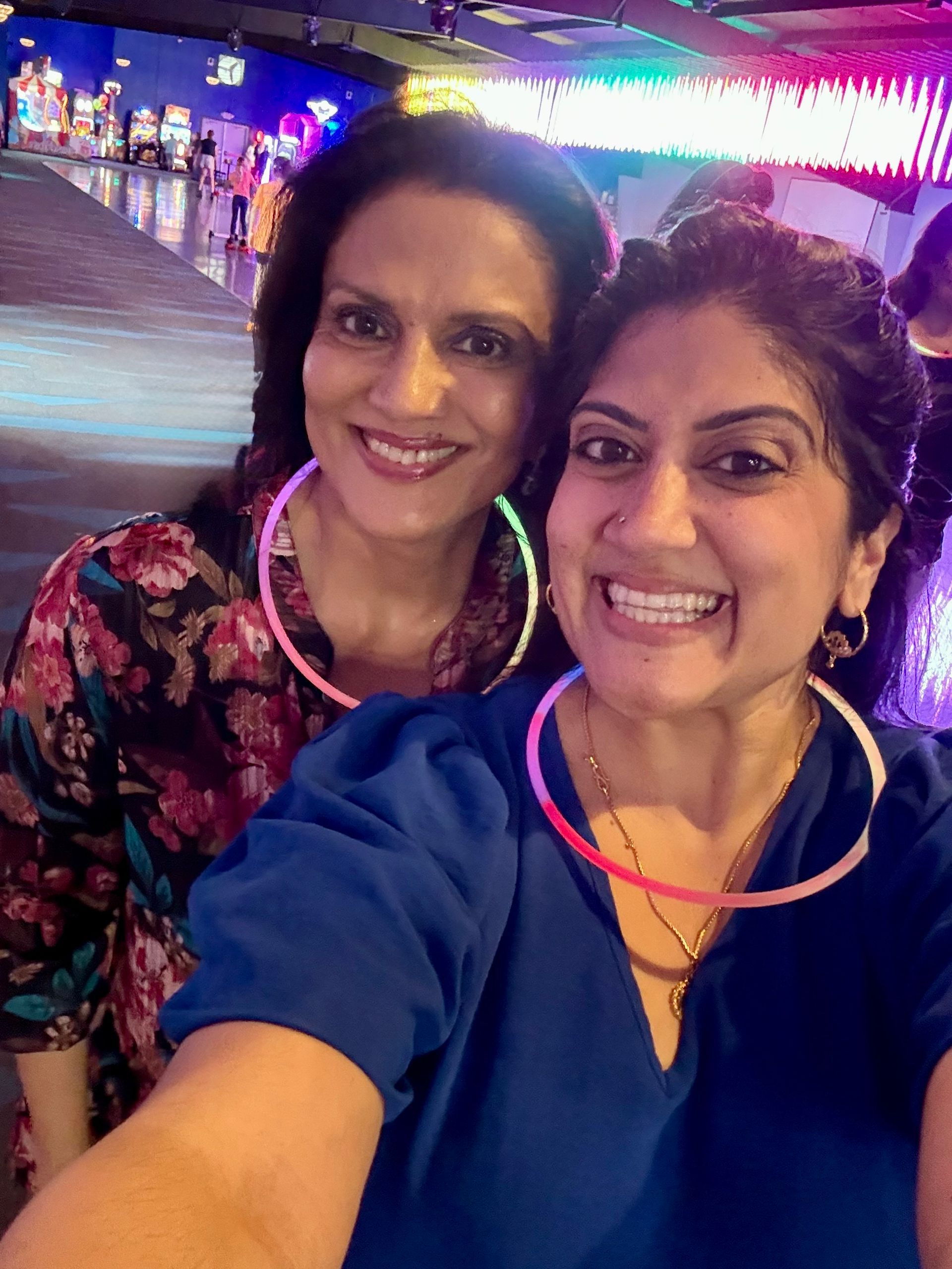 Two women smiling, wearing glow stick necklaces, in a brightly lit arcade.