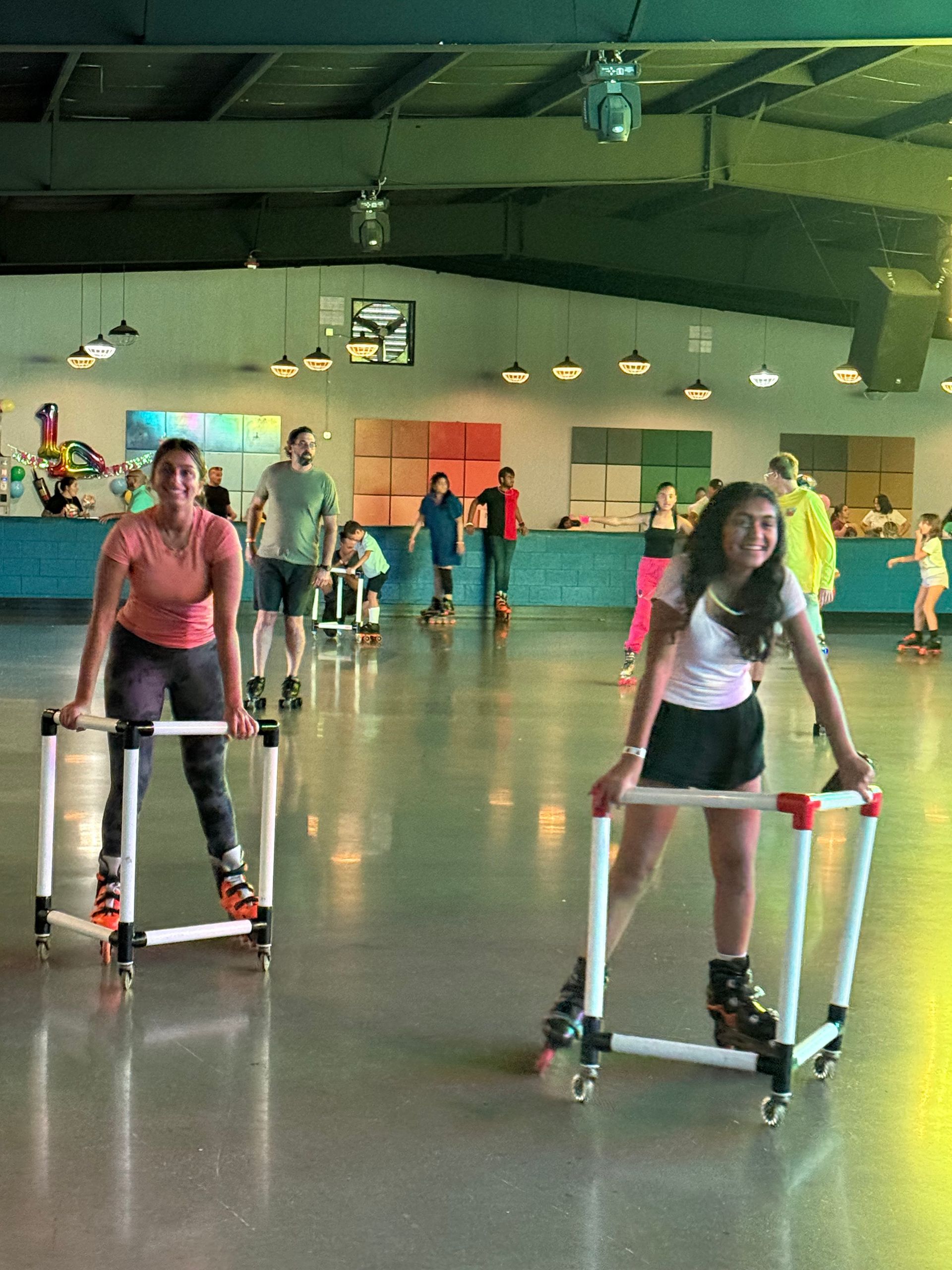 Two people roller skate, using white frames for balance, in an indoor rink with others skating.