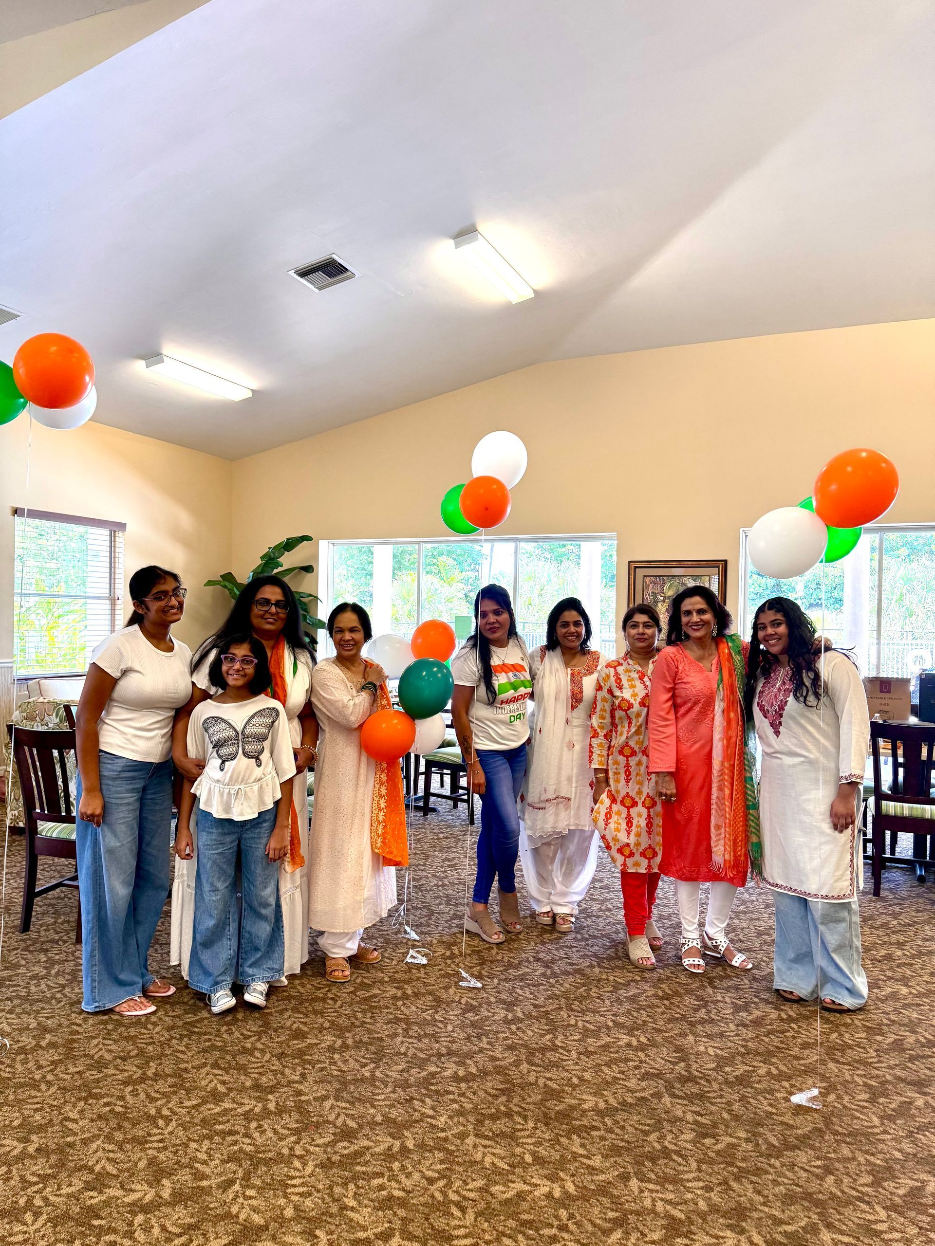 Group of people, mostly women, in colorful attire with balloons, celebrating in a room with windows and lights.