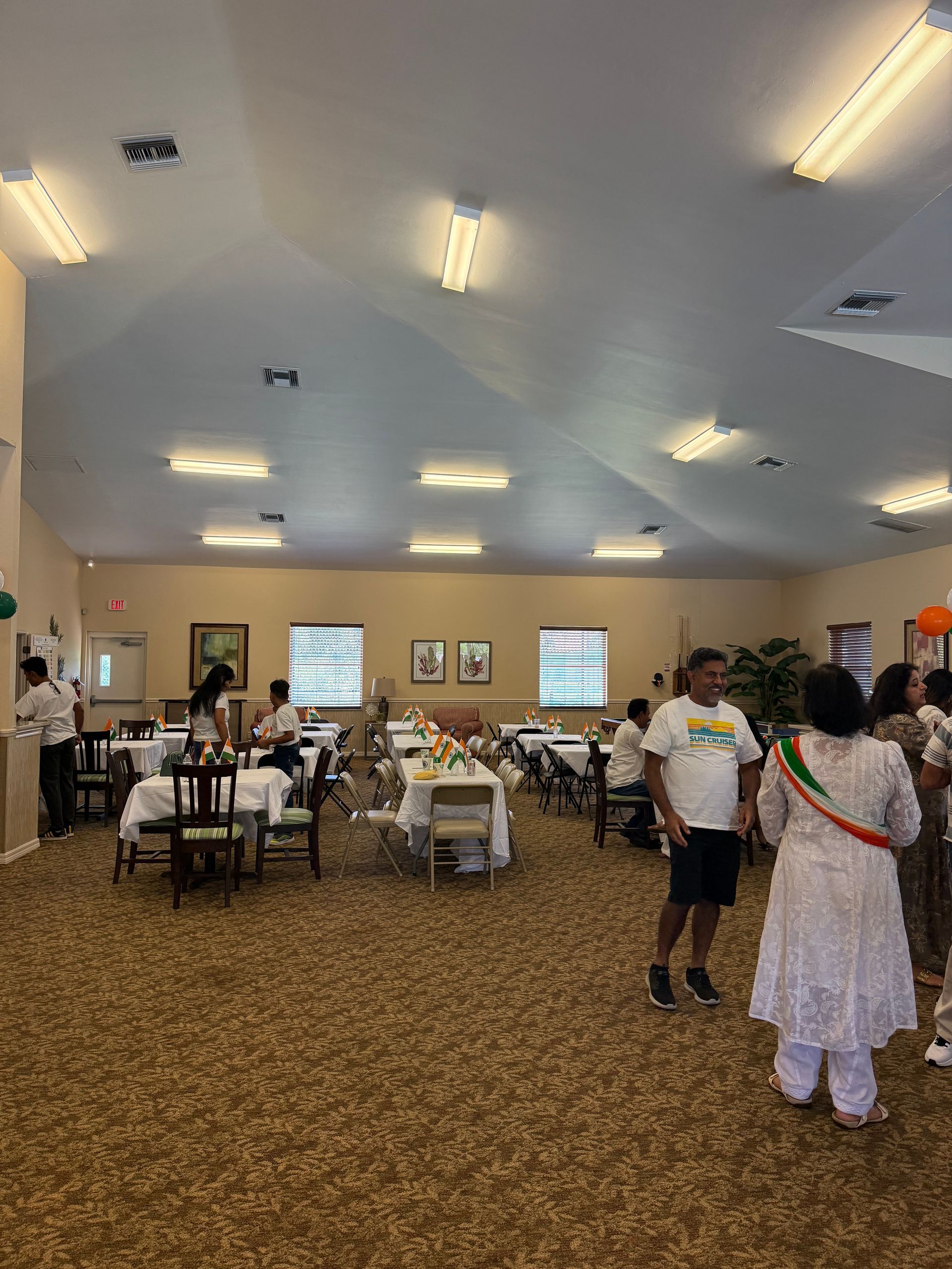 People at an event in a light-filled room with tables. Some are seated, some are standing, and decorated tables are set.