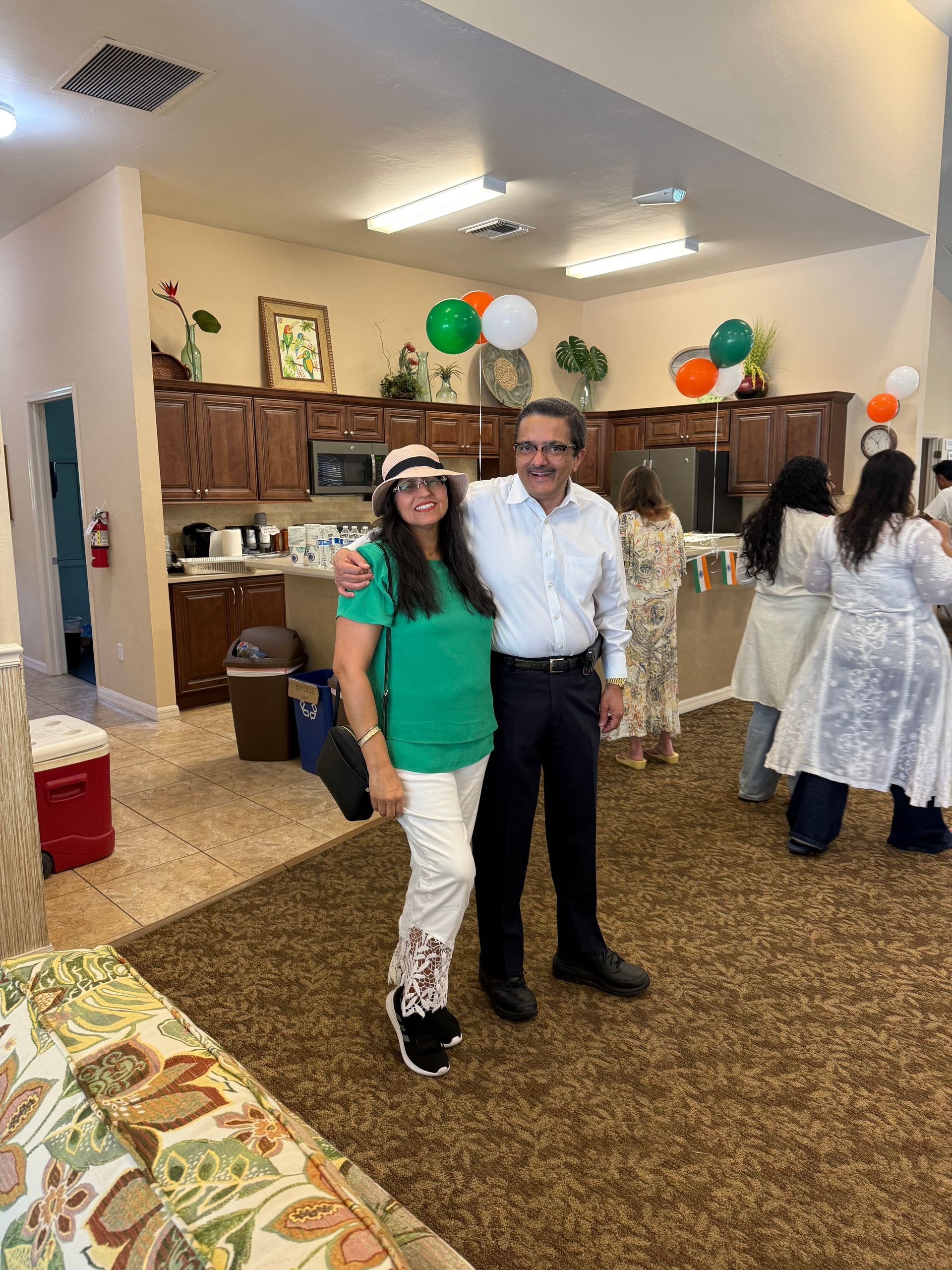 A man and woman in festive attire pose indoors; green and orange balloons in background.