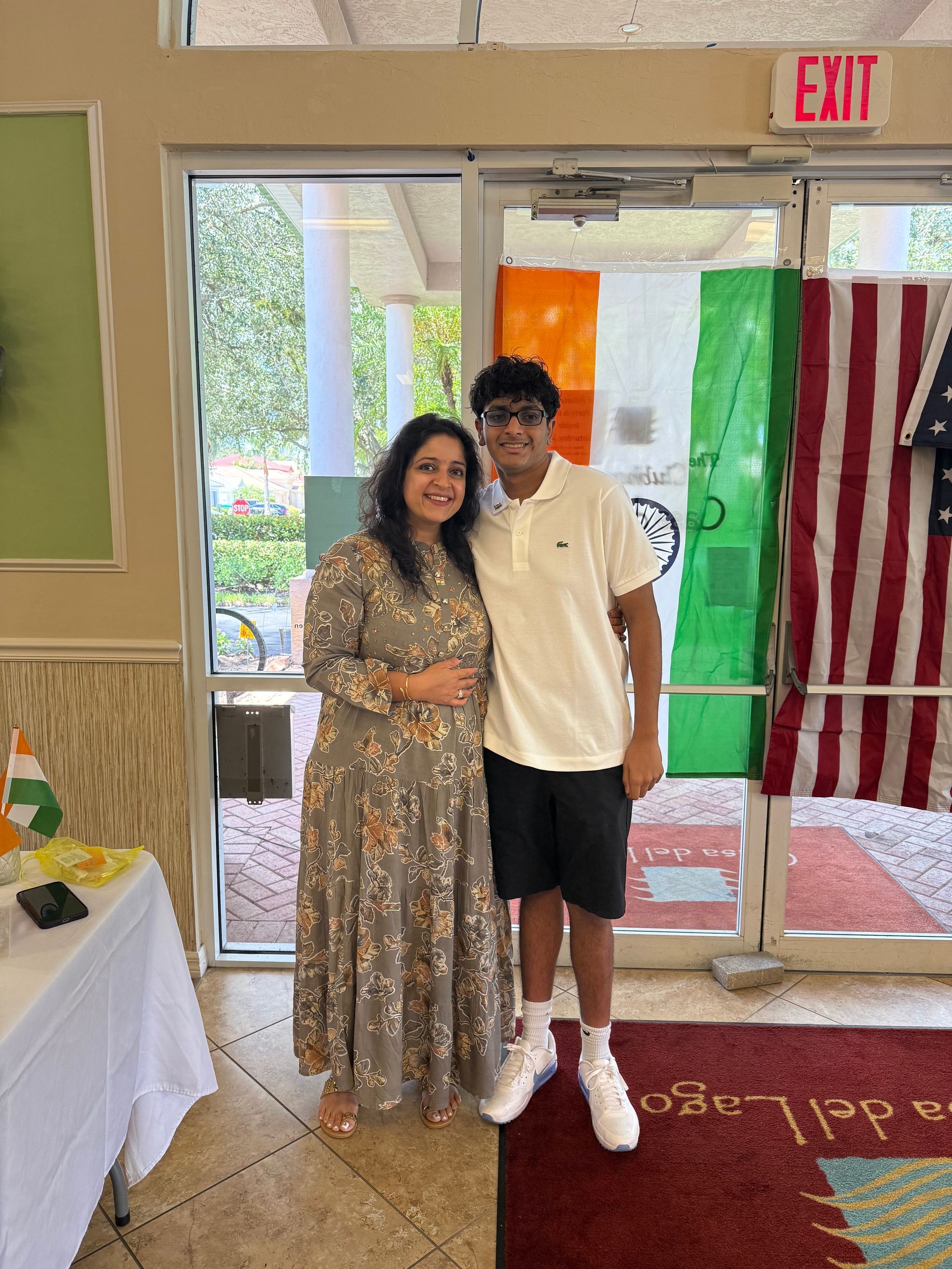 Woman and boy stand inside near Indian and American flags. Woman in floral dress smiles. Boy in polo, shorts smiles.