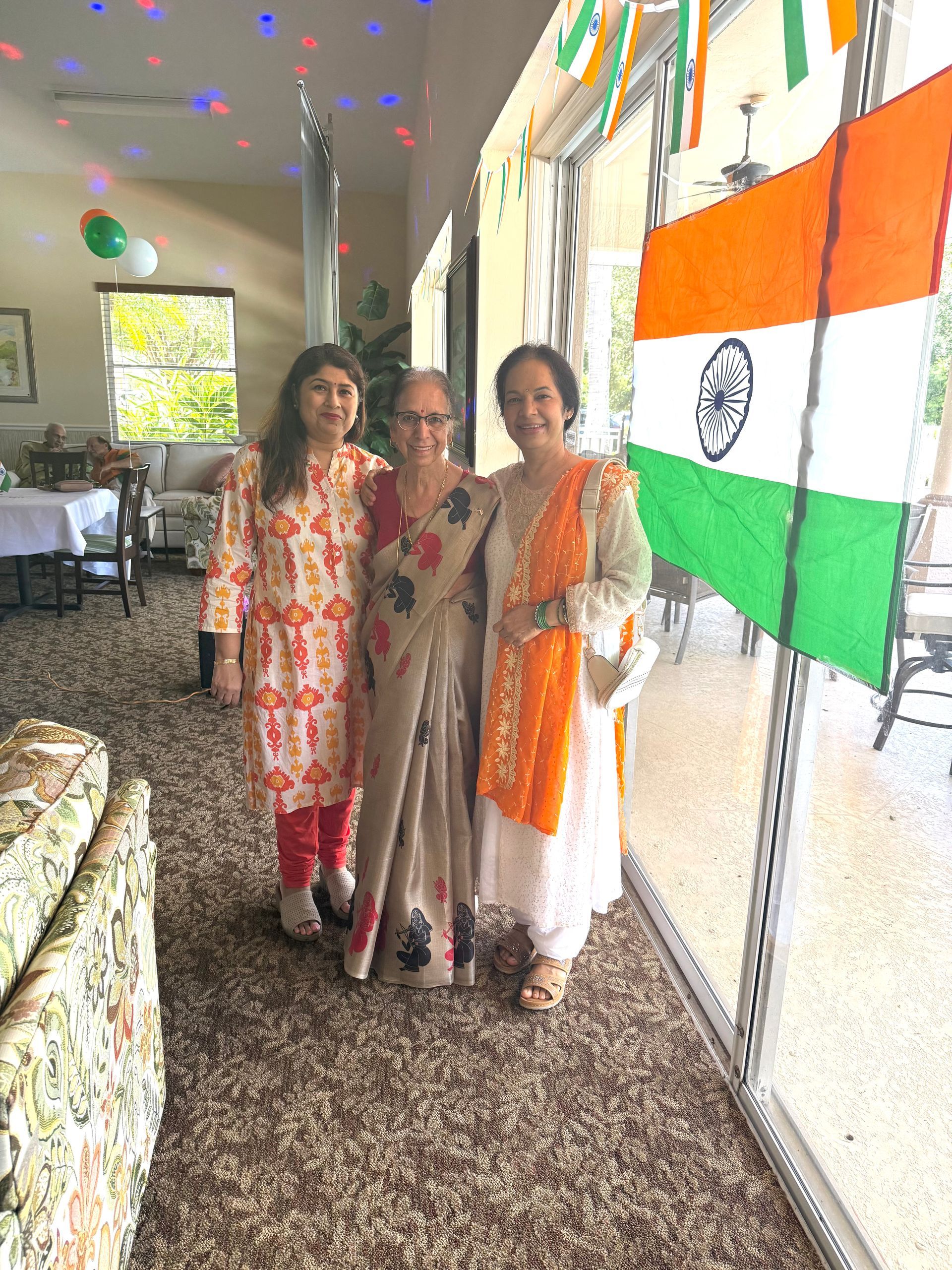Three women pose for a photo with an Indian flag in a decorated room.