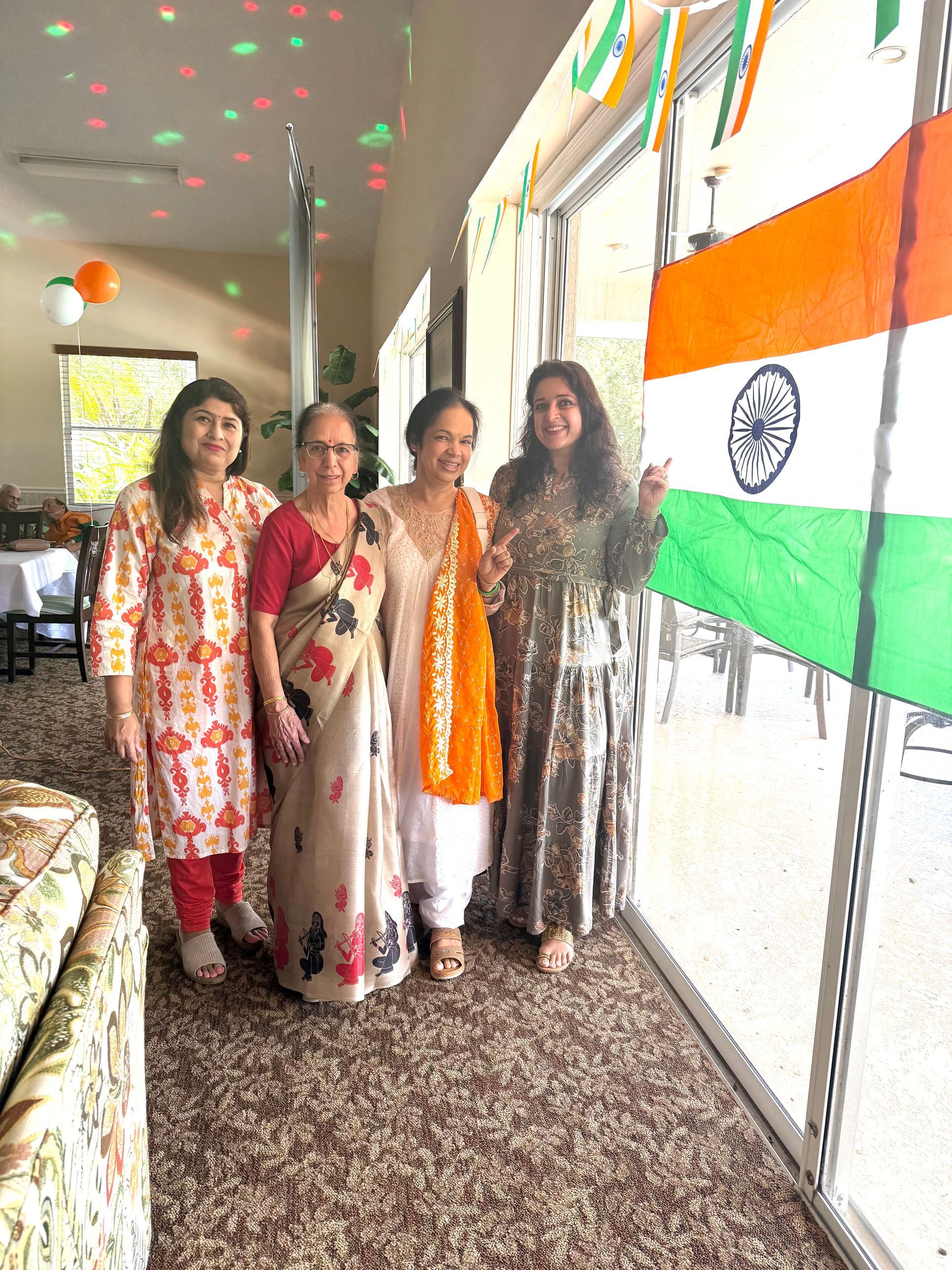 Four women pose with an Indian flag; interior with green and orange decorations.