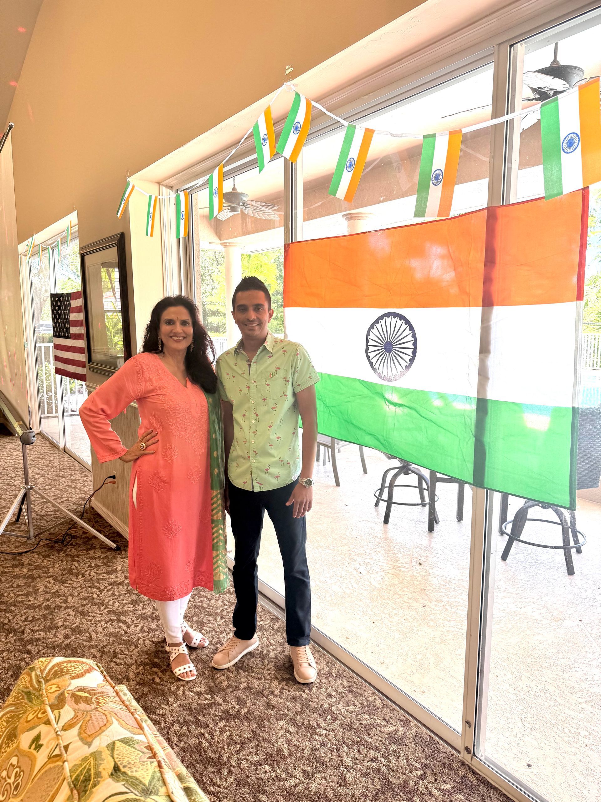 Two people pose next to an Indian flag. Woman wears pink, man wears green; a windowed room.