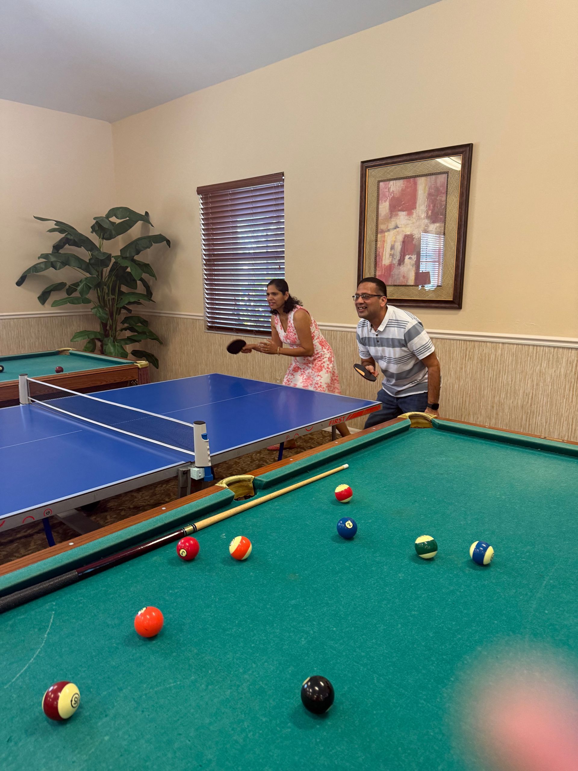 People playing ping pong and billiards in a game room; woman serving, man waiting.