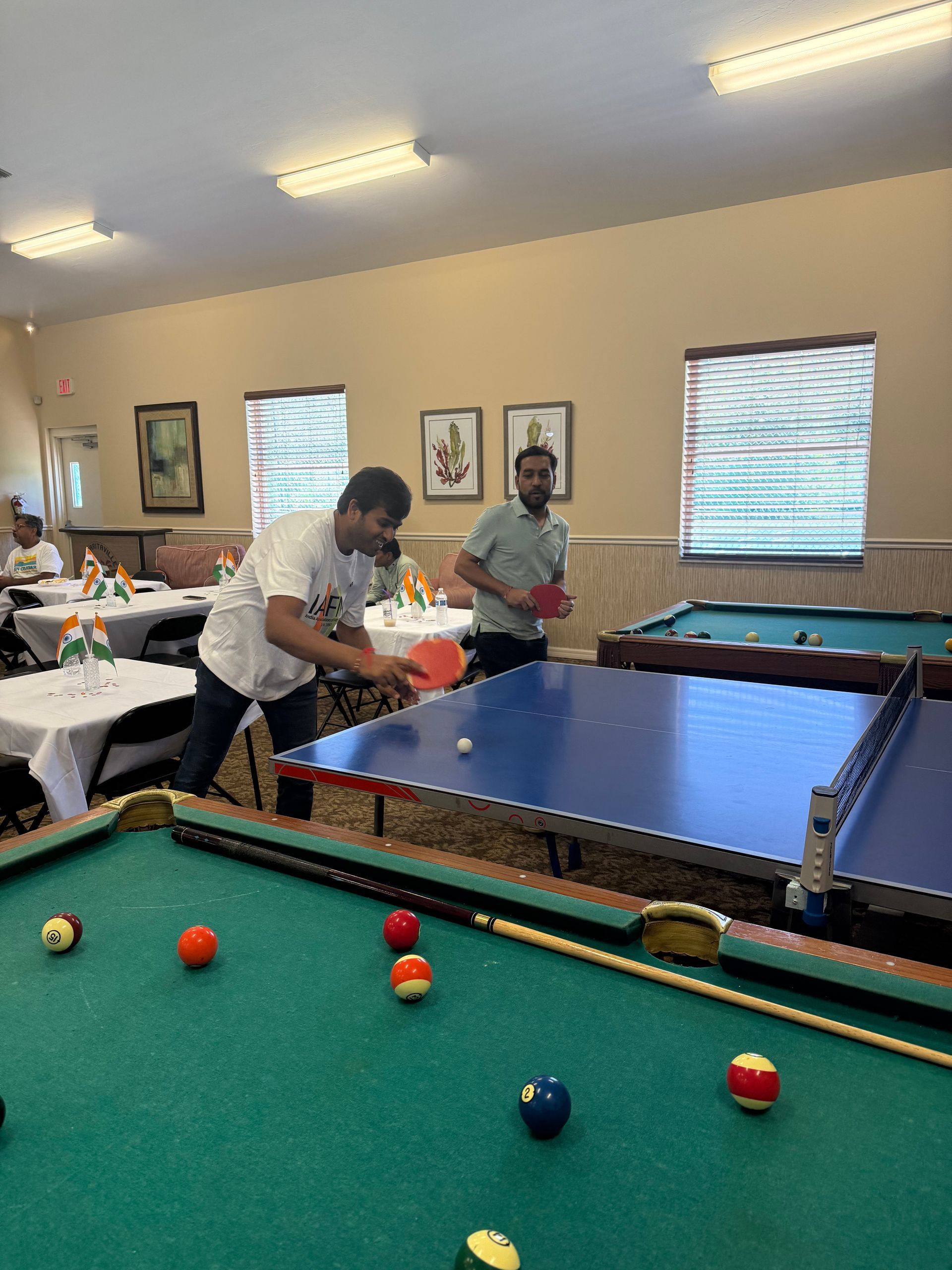 Two men playing ping pong in a room with pool tables. One hits the ball with a paddle.