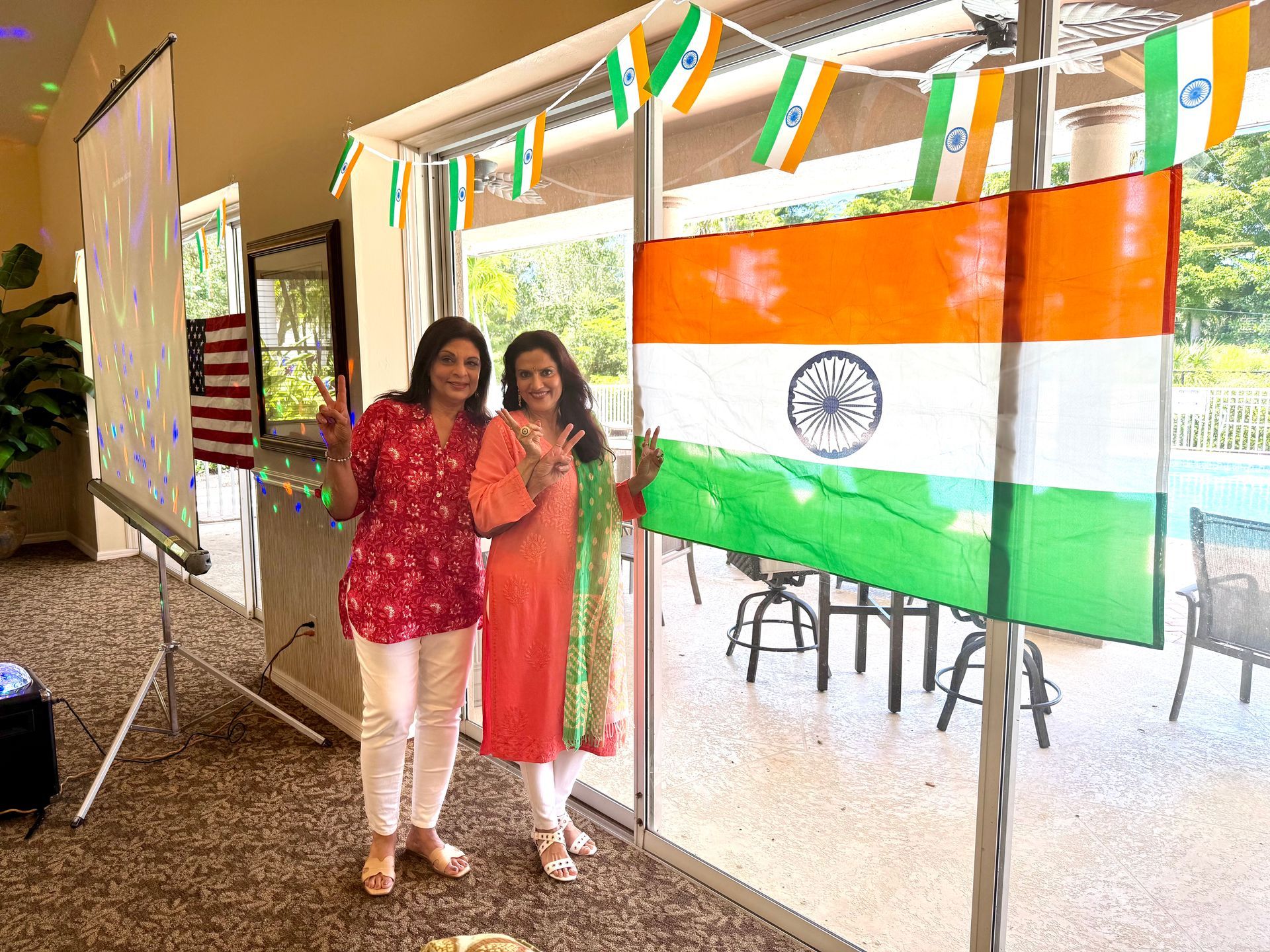 Two women pose with India's flag, celebrating in an indoor/outdoor setting. Flags decorate.