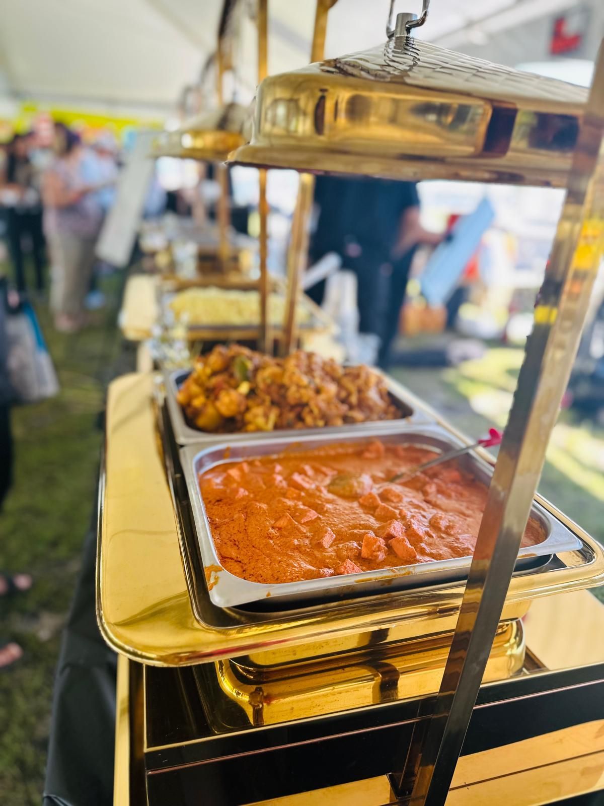 Buffet with golden serving dishes, containing curry and other Indian food, at an outdoor event.