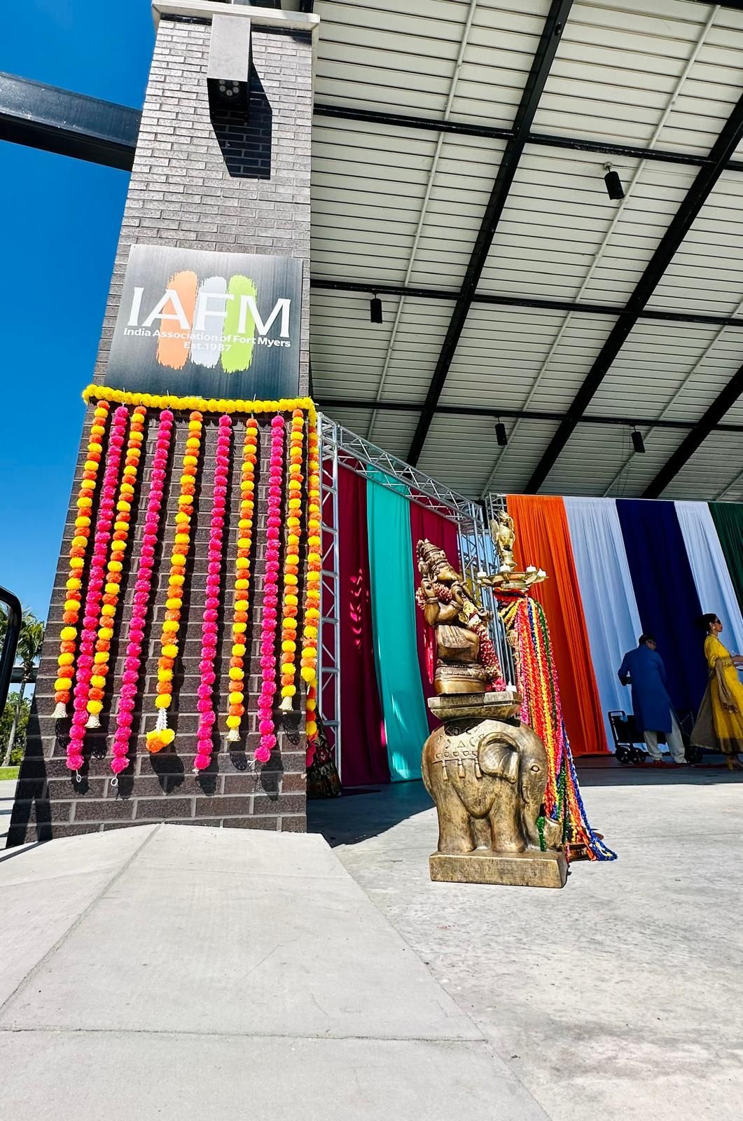 Decorated pillar with flower garlands and colorful fabrics, a golden statue, and people in traditional clothing outside.