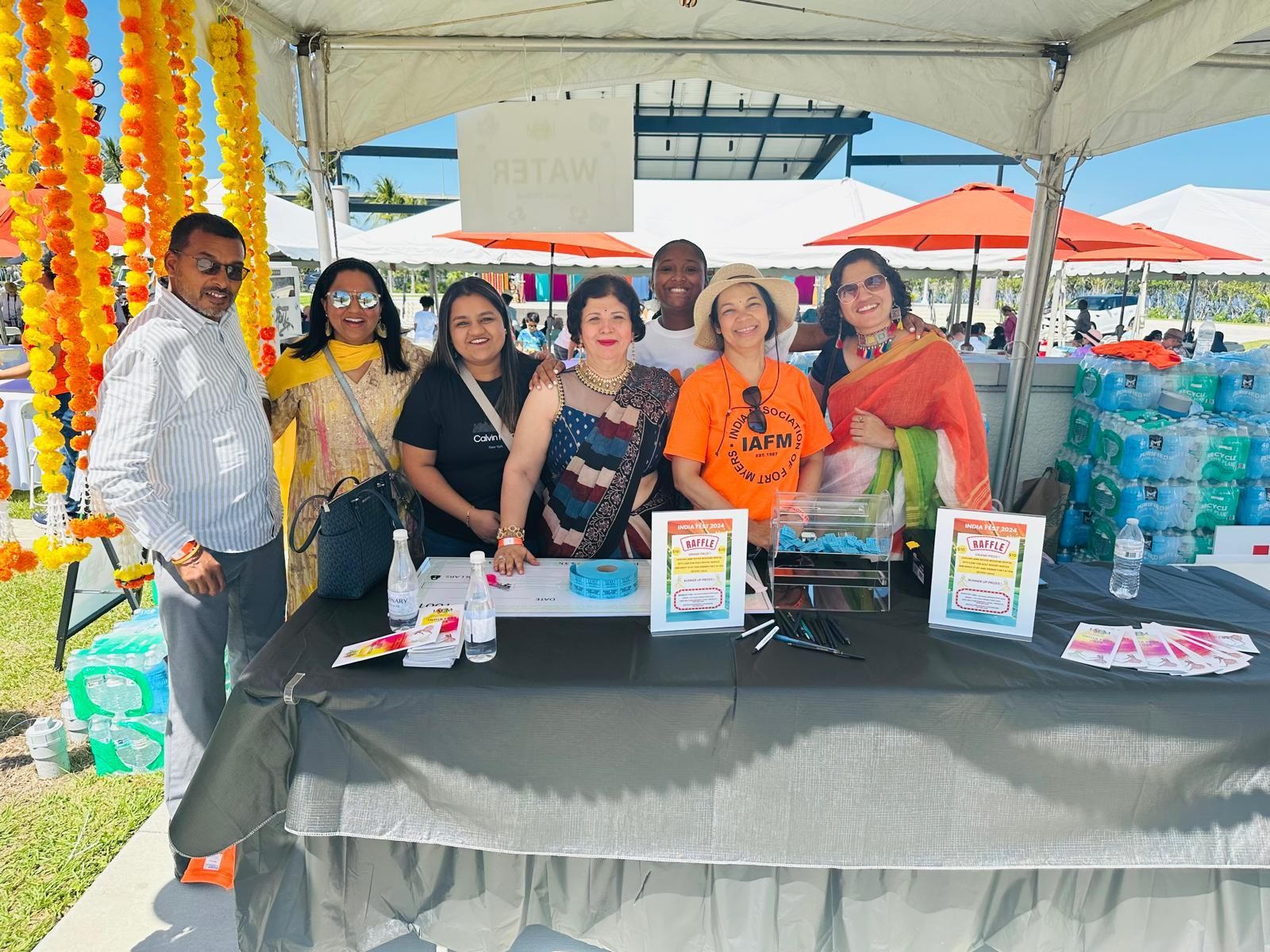 Group of people smiling at a booth under a tent, outdoors. Tables with items on display.