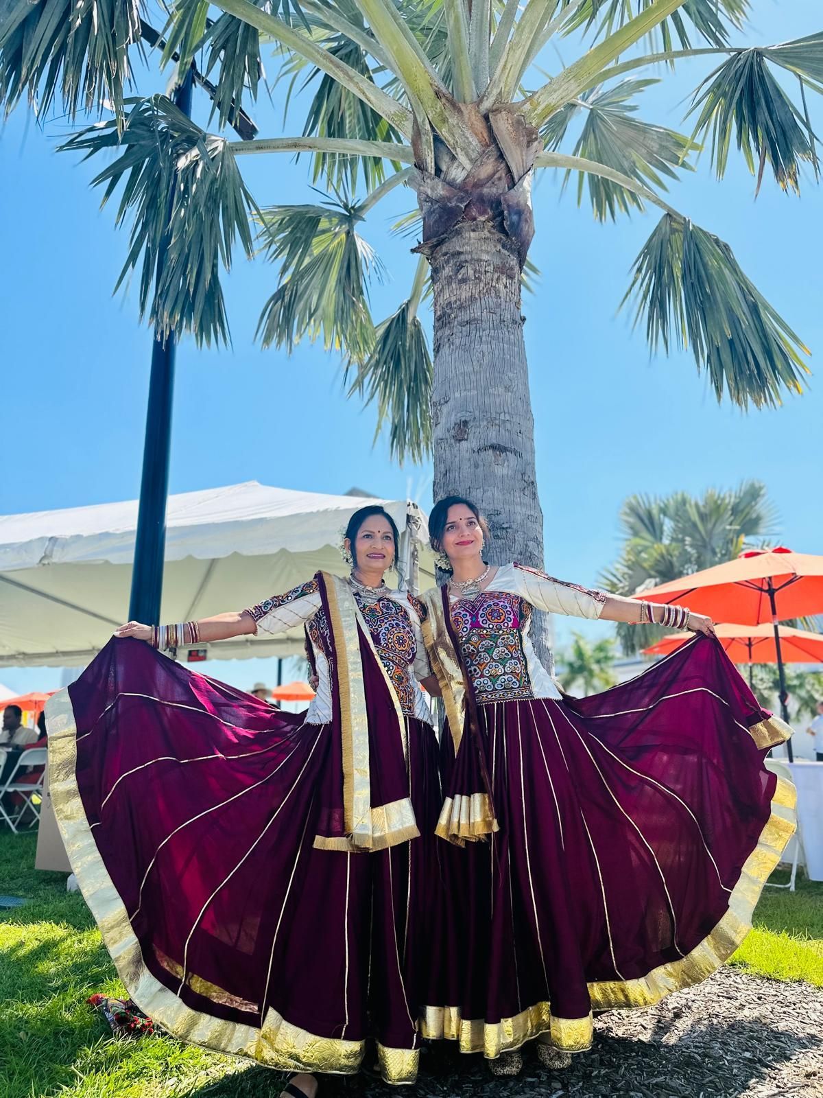 Two women in maroon and gold Indian dresses pose outdoors under a palm tree.