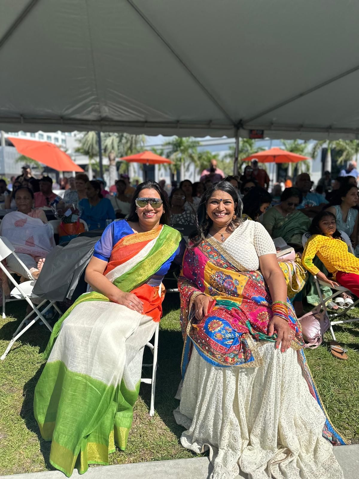 Two women in saris smile outdoors under a tent, surrounded by a crowd.
