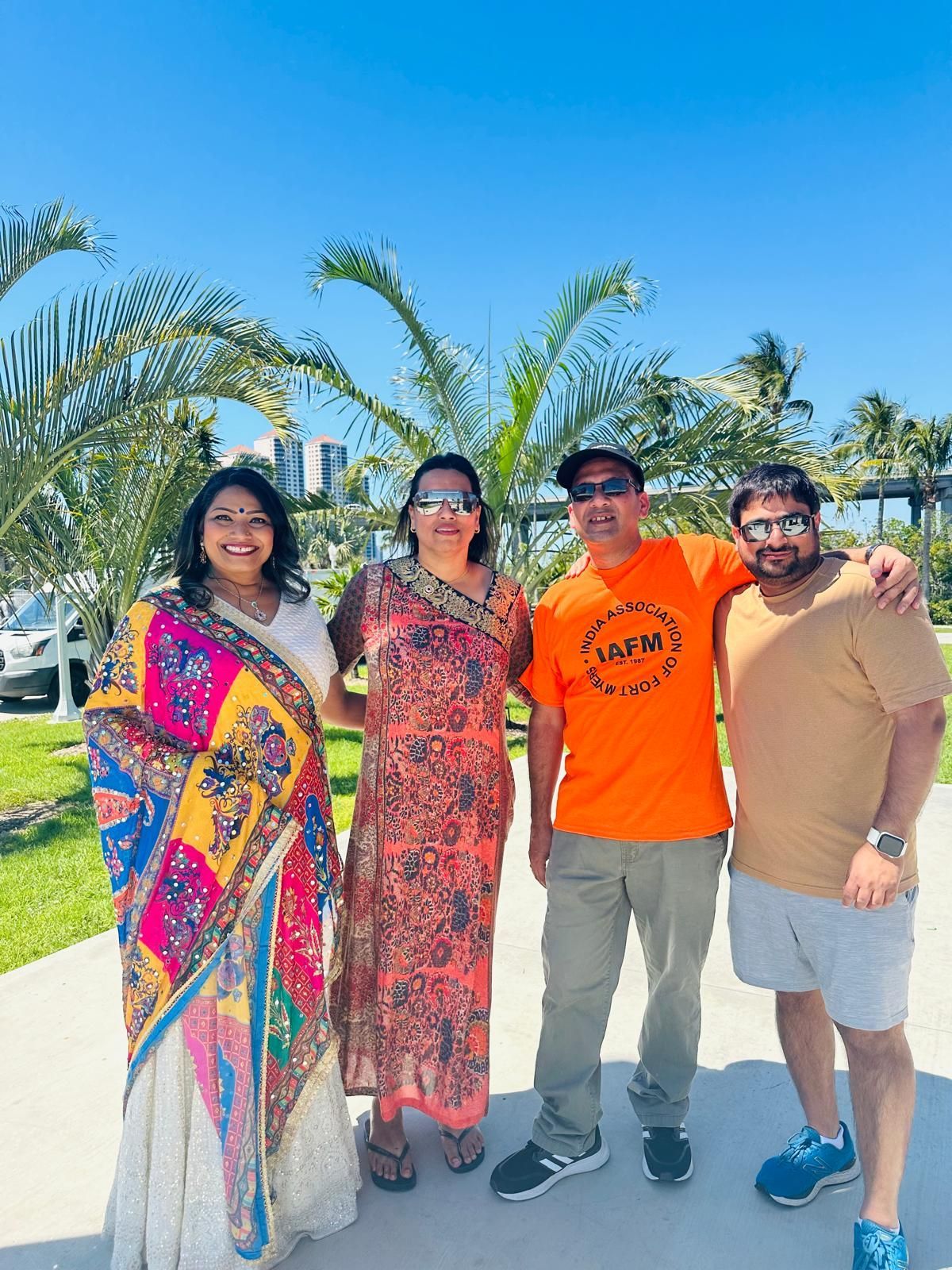 Four people smiling outdoors. Two women in vibrant dresses, two men in casual wear, palm trees.