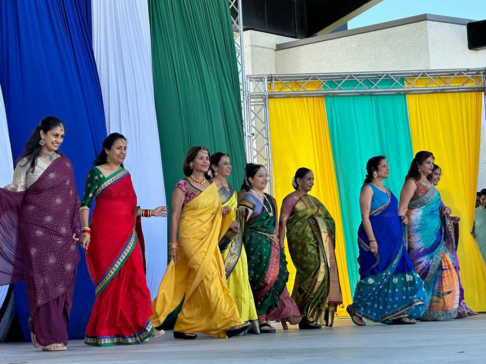 Group of women in colorful saris on a stage, smiling and walking forward in an outdoor setting.